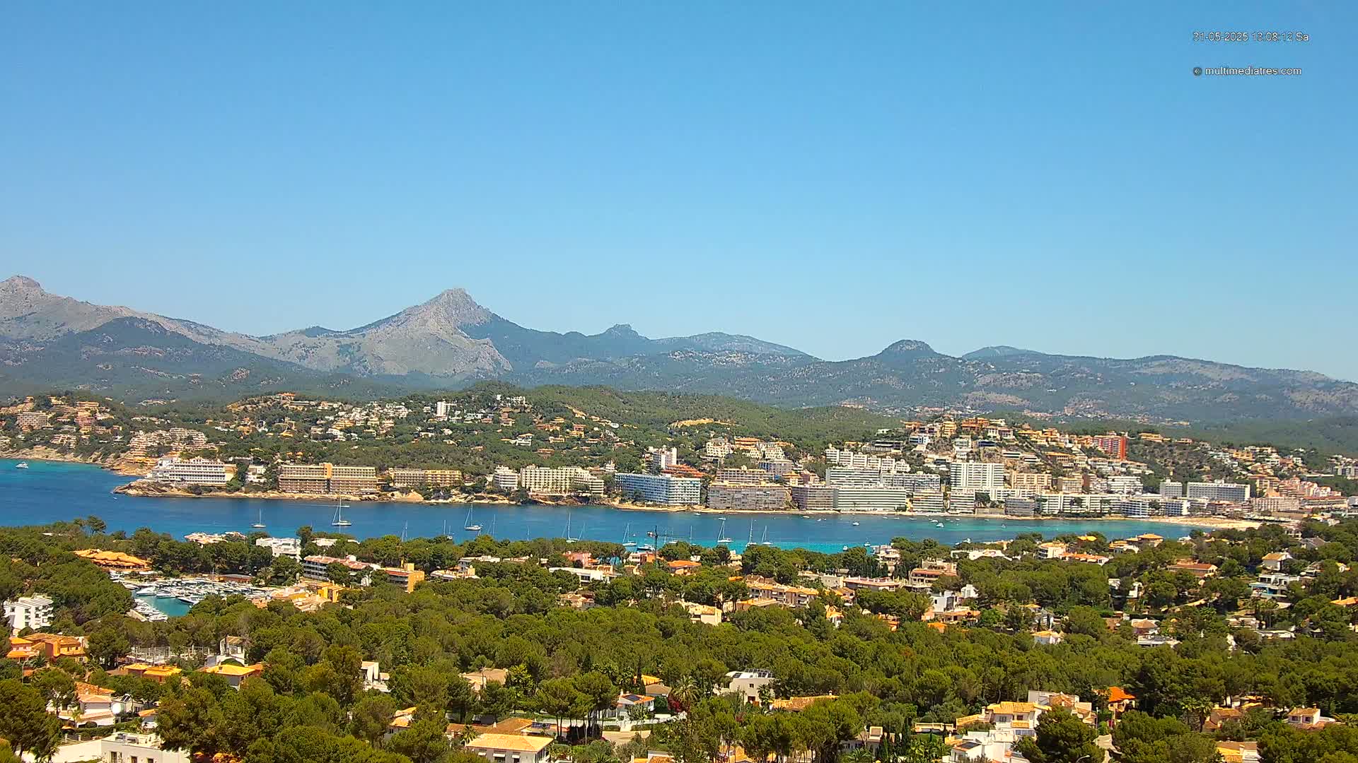 A coastal town nestled in a verdant valley, with a clear blue bay and a mountain range in the background under a sunny sky.