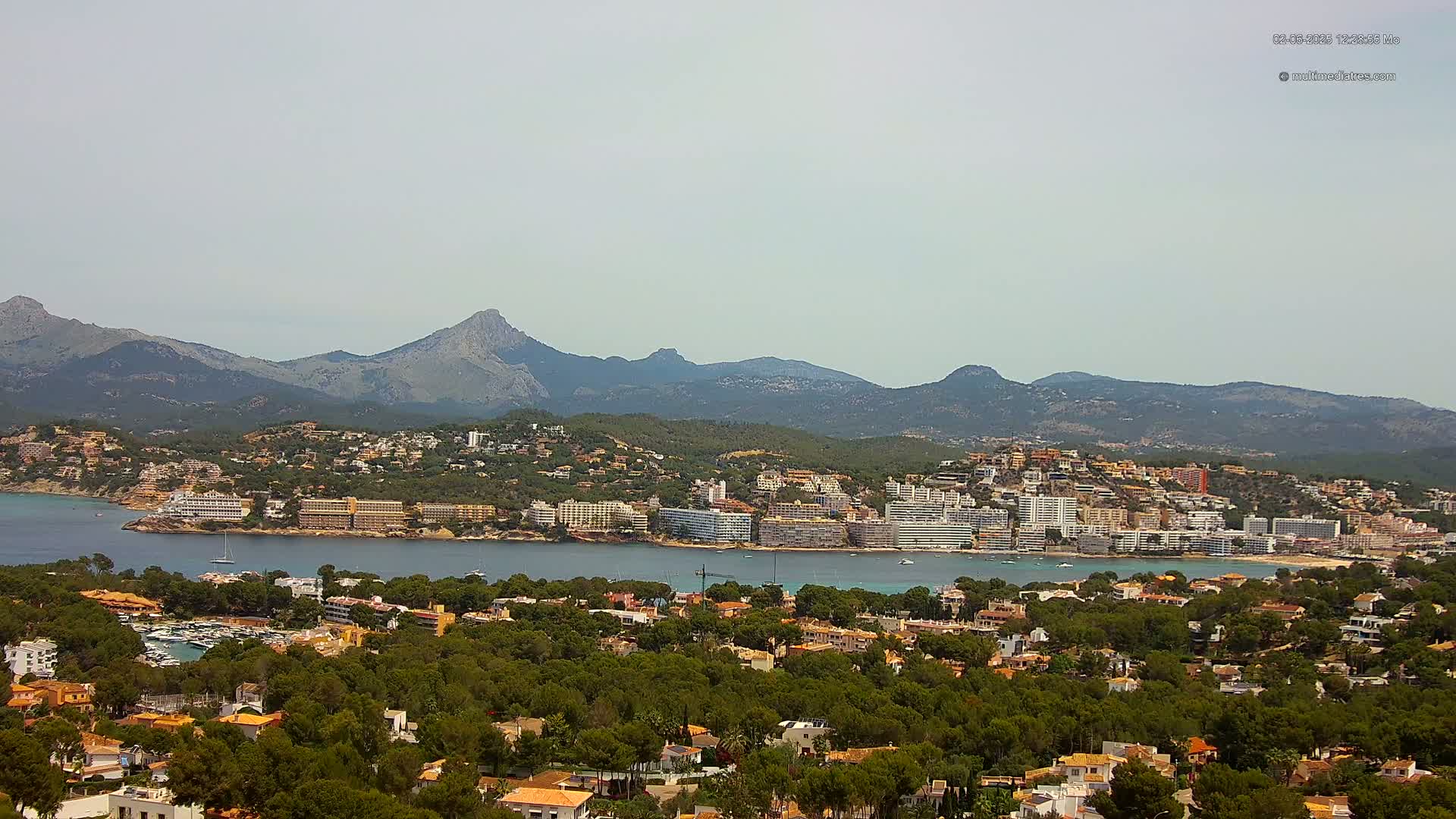 A coastal town nestled between a calm bay and a mountain range under a hazy sky.