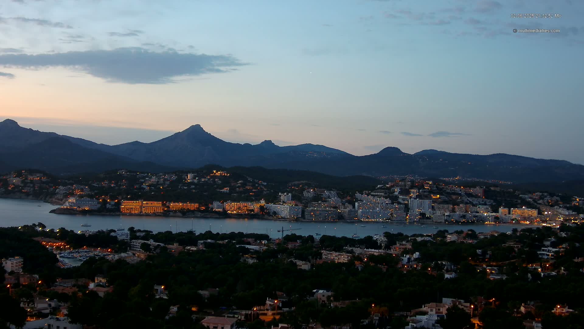 A coastal town with illuminated buildings nestled at the base of dark mountains under a twilight sky.