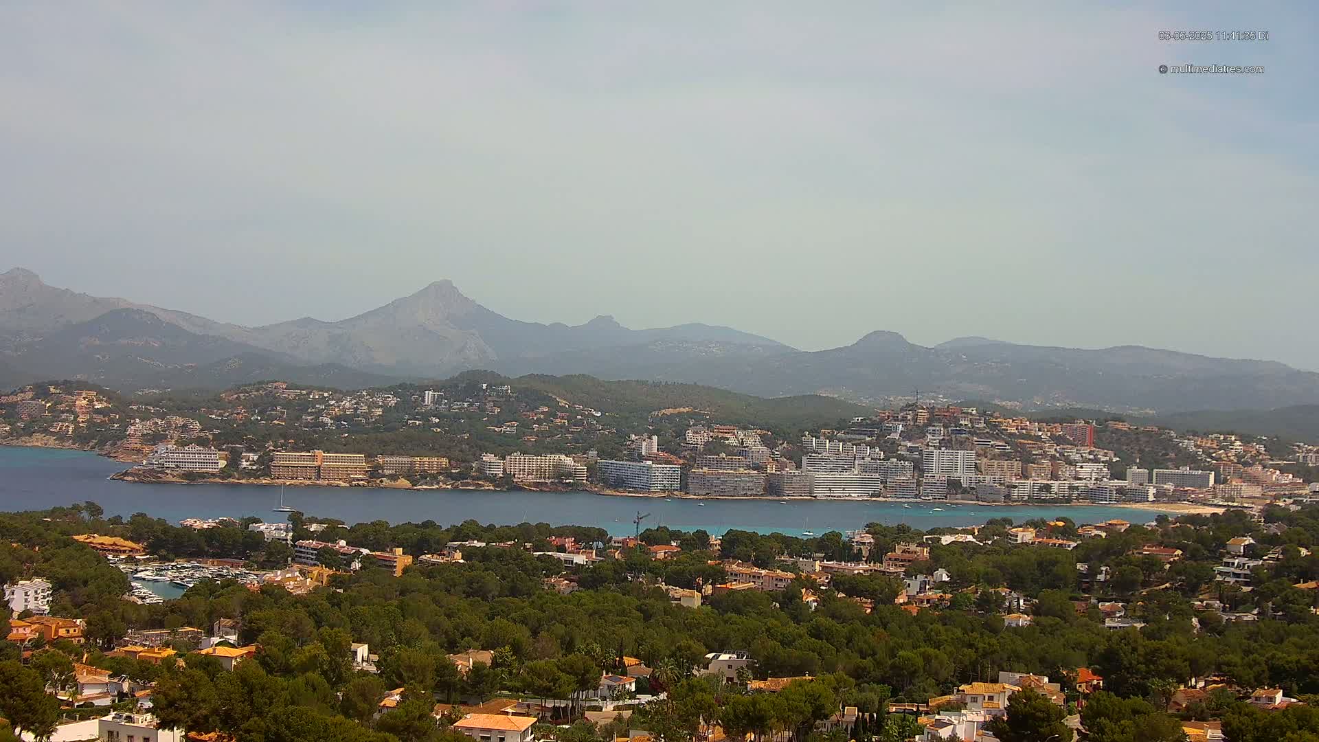 A coastal town nestled amongst green hills and a calm bay, with hazy mountains in the background under a light overcast sky.