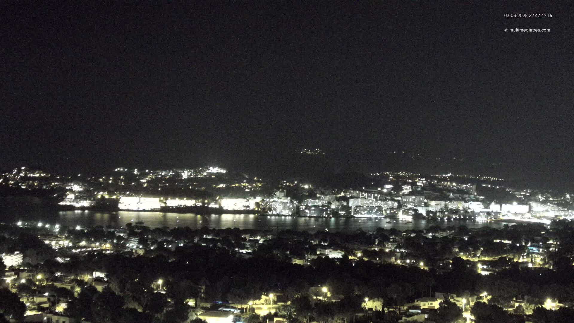 A nighttime, high-angle view shows a coastal city illuminated with lights across a body of dark water, under a clear, dark sky.