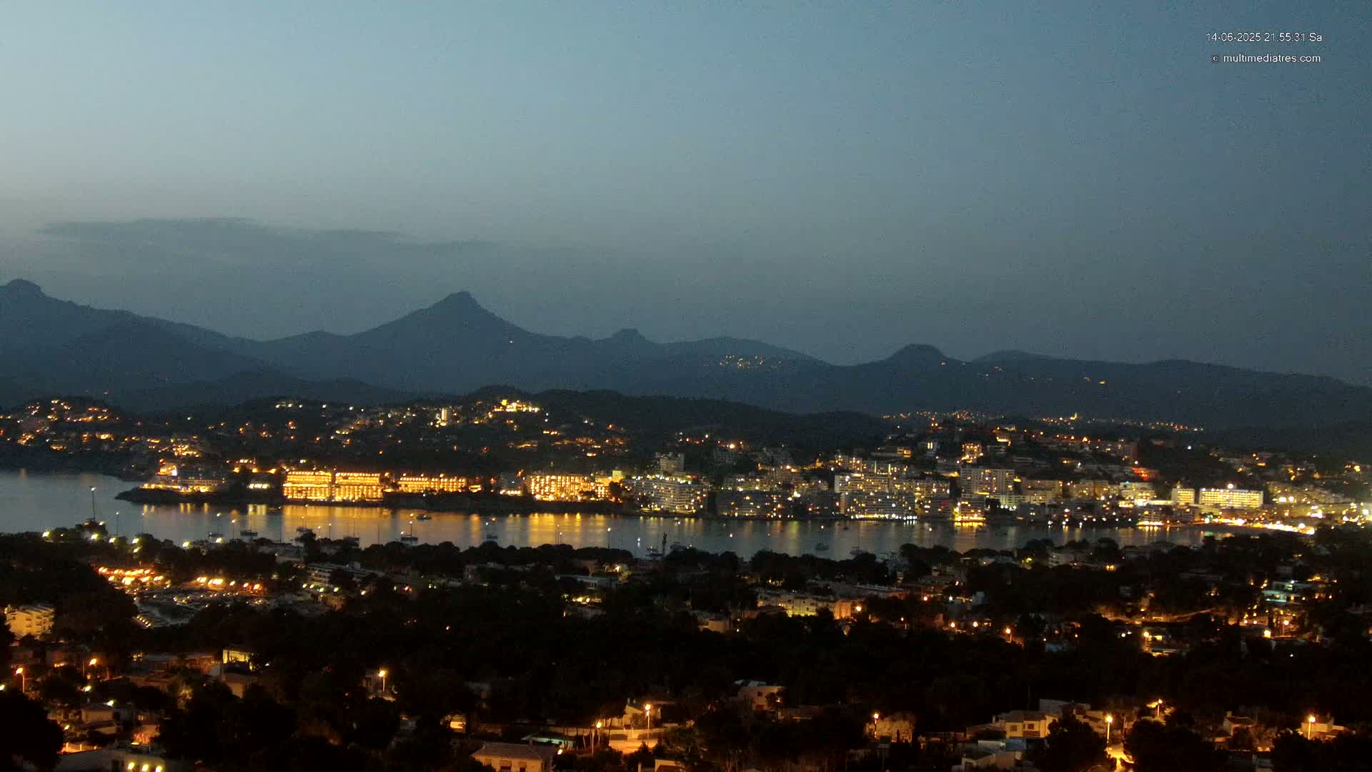 A nighttime, high-angle view shows a coastal city illuminated with lights across a body of dark water, under a clear, dark sky.