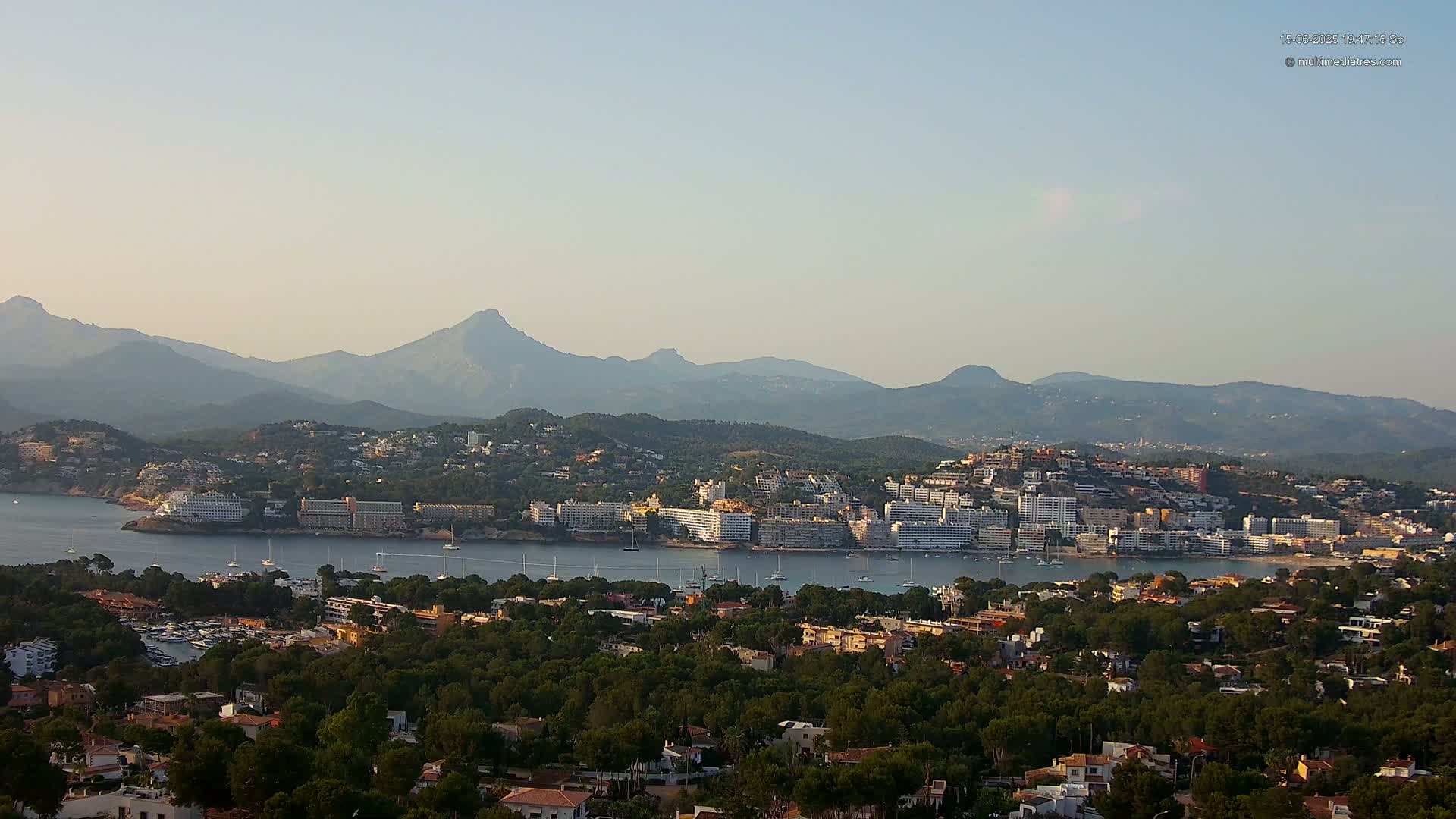 A coastal town nestled among green hills and mountains under a clear, light-blue sky.