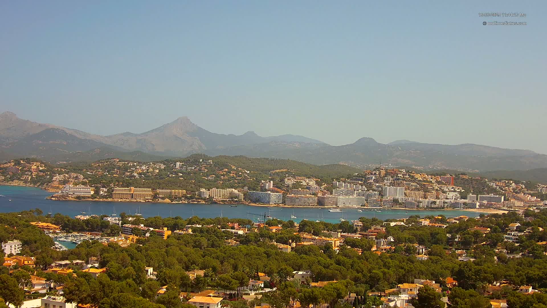 A coastal town nestled between a calm bay and a mountain range under a clear, sunny sky.
