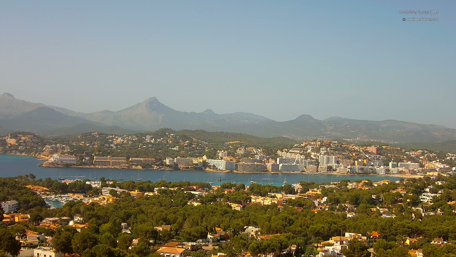 A coastal town nestled amongst green hills and mountains under a hazy, sunny sky, with a calm bay filled with boats.