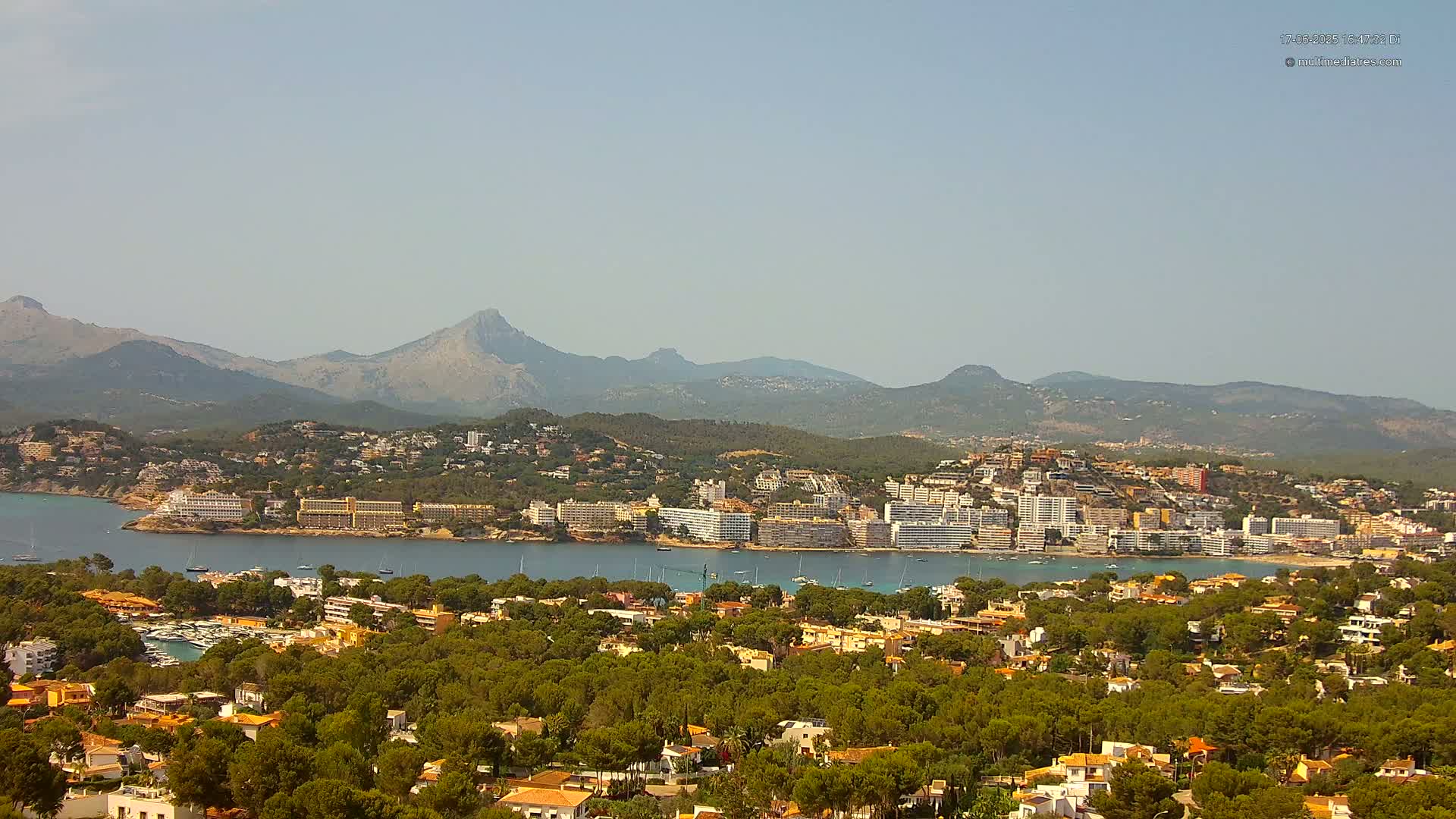 A coastal town nestled amongst green hills and mountains under a hazy sky.