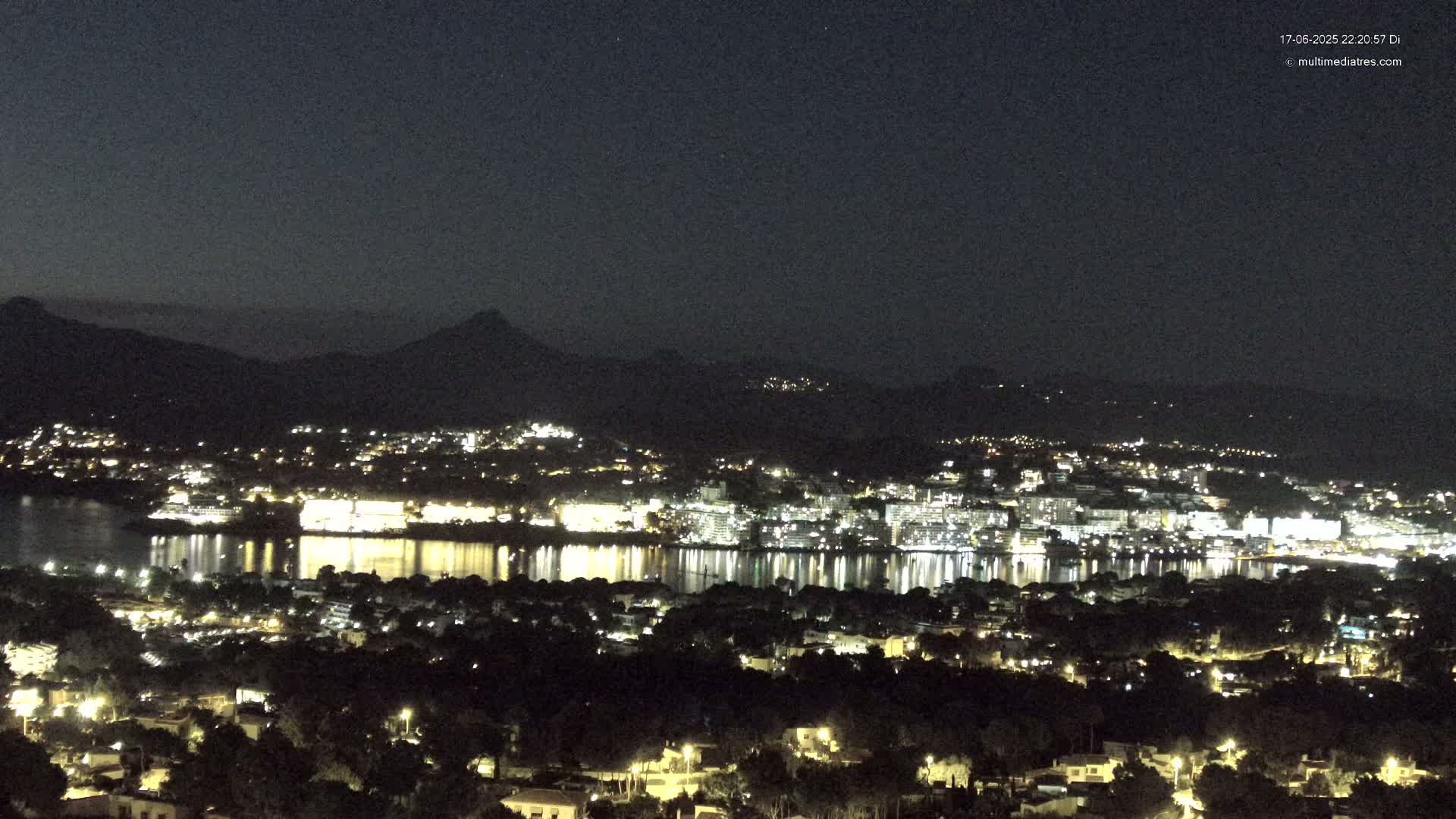 A nighttime view of a coastal town illuminated with lights, nestled against a dark mountain range under a clear, starry sky.