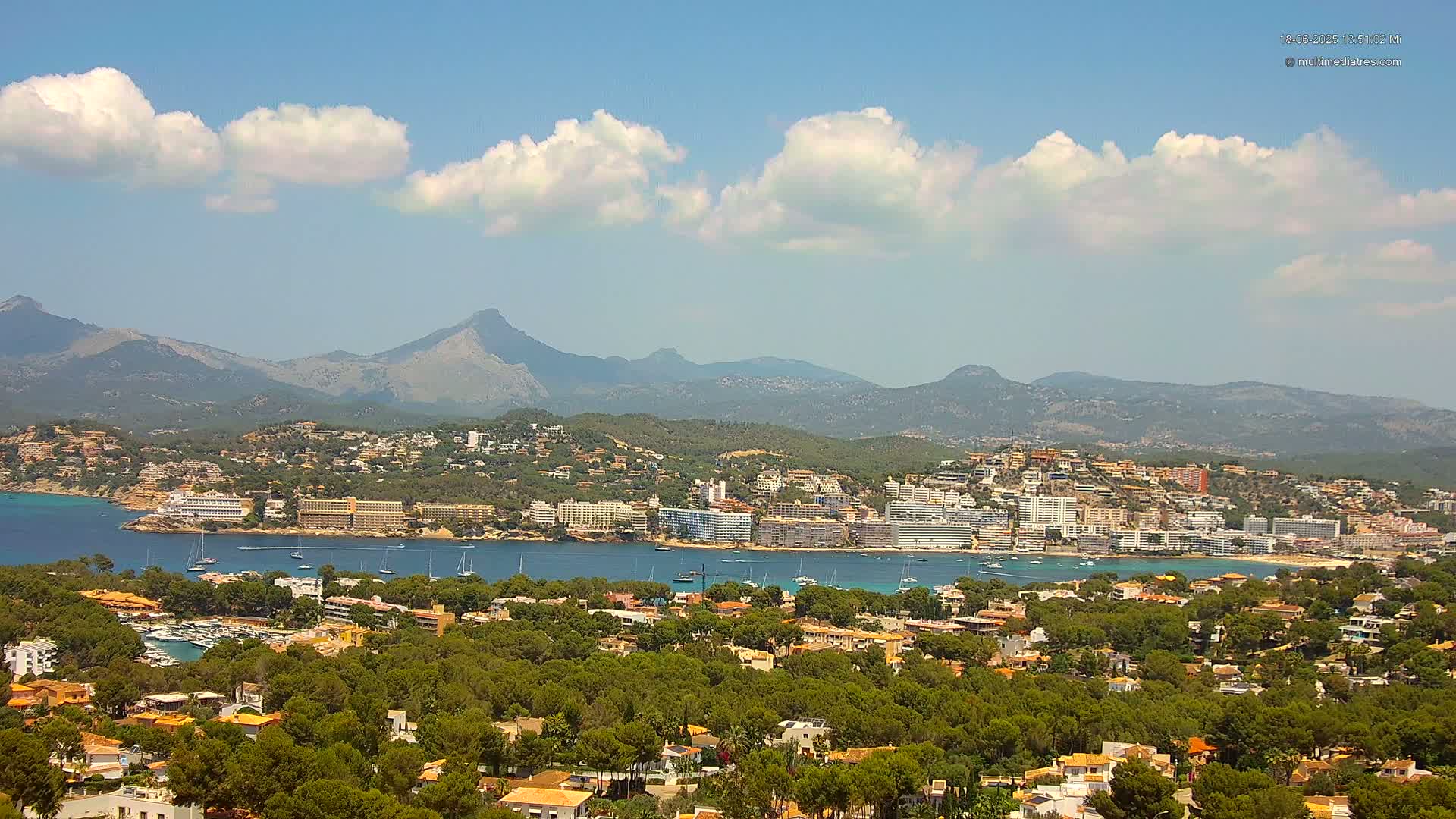 A coastal town nestled in a verdant valley, with a calm bay and mountains in the background under a mostly sunny sky with scattered cumulus clouds.