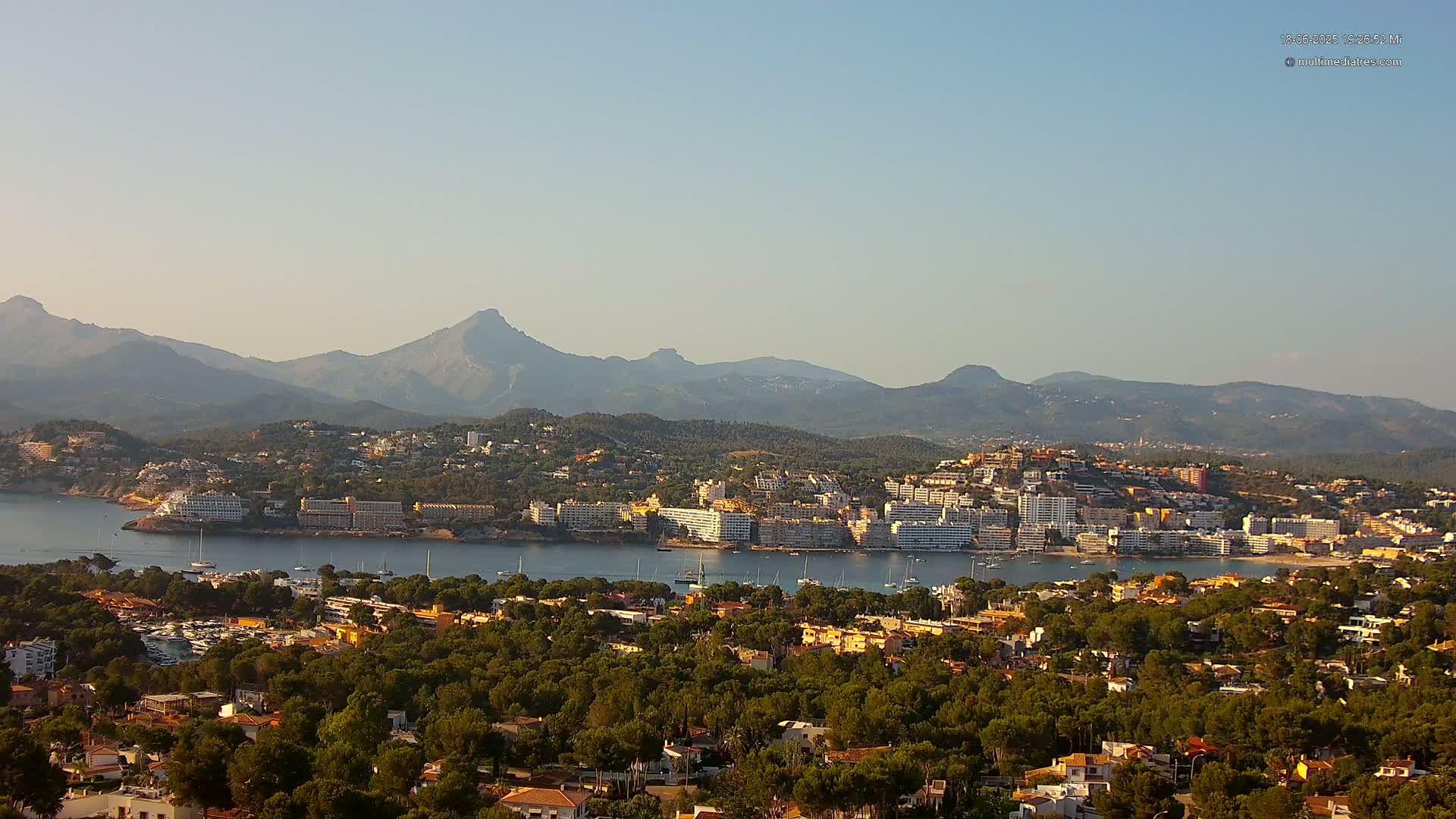A coastal town nestled in a bay, surrounded by lush green hills and mountains under a clear, light-blue sky.