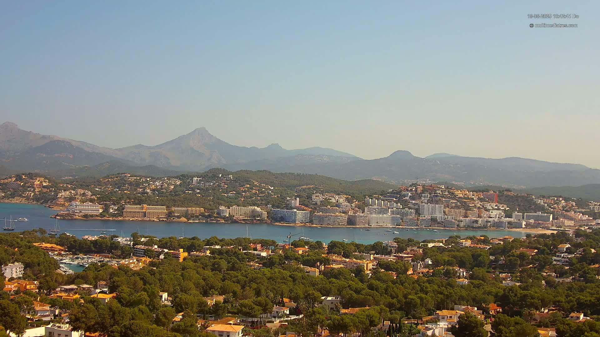A coastal town nestled in a bay, surrounded by lush green hills and mountains under a clear, sunny sky.