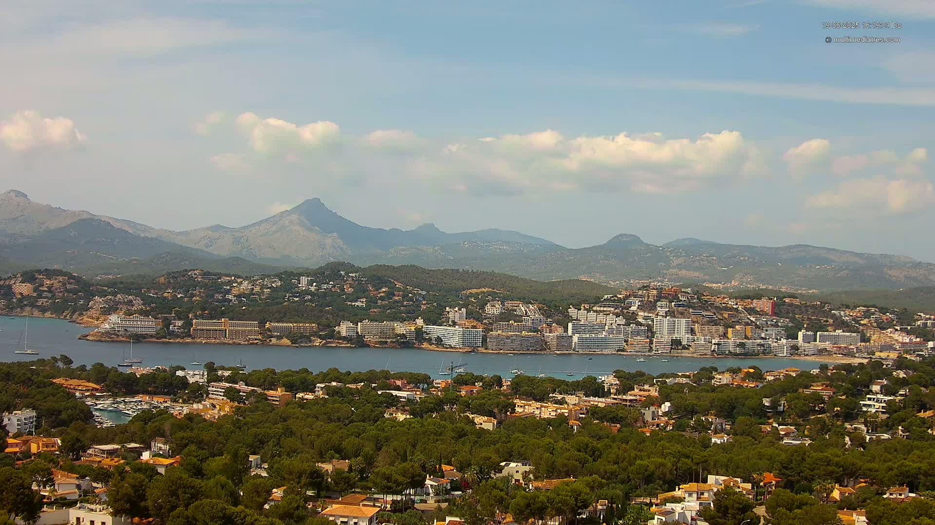 A coastal town nestled among green hills and mountains under a partly cloudy sky.