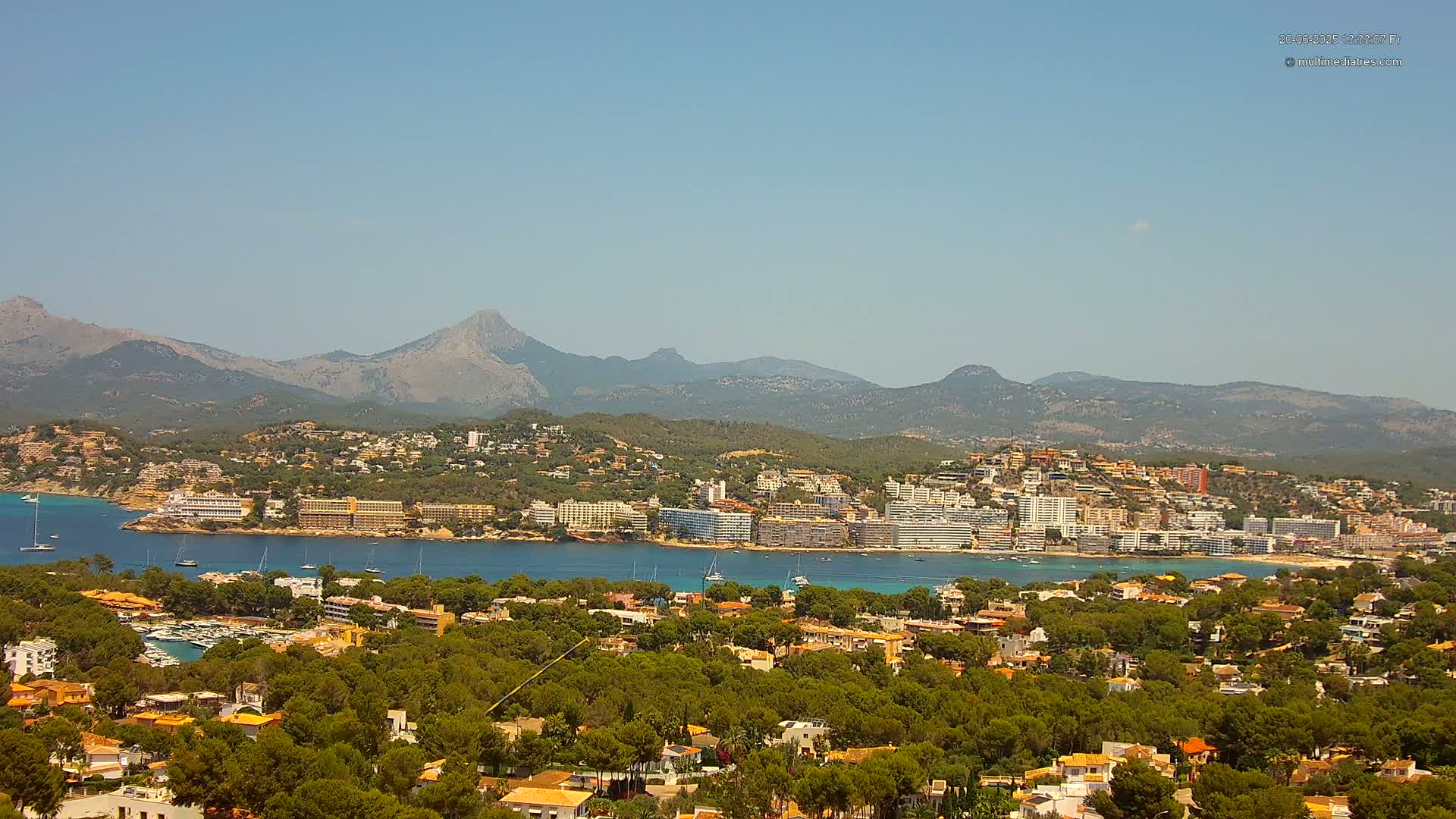 A coastal town nestled among green hills and a clear blue bay, backed by a range of mountains under a sunny sky.