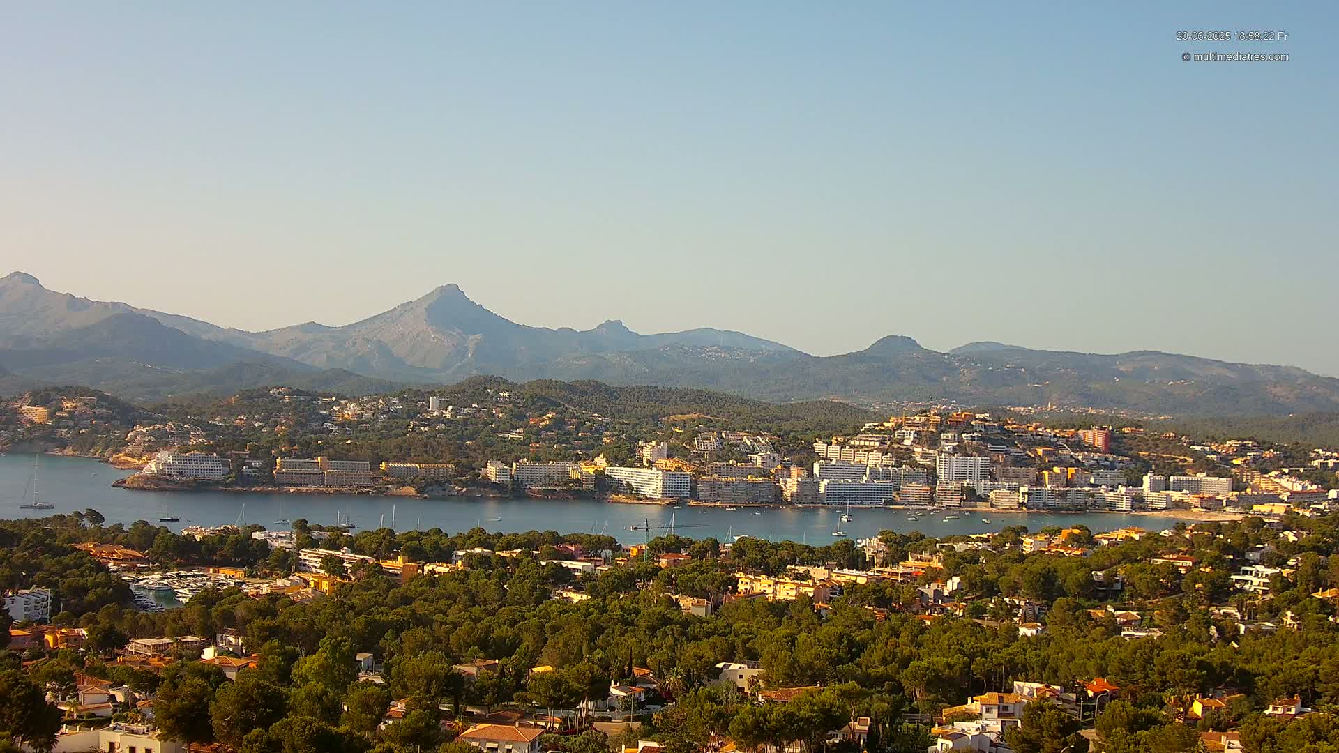 A coastal town nestled among green hills and mountains, under a clear, bright sky, with a calm body of water separating the town from a peninsula.