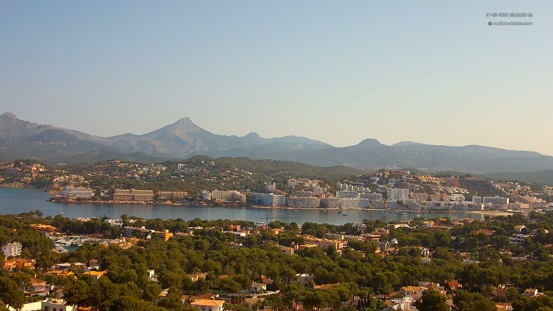 A coastal town nestled amongst green hills and a clear blue bay, with mountains in the background under a hazy sky.