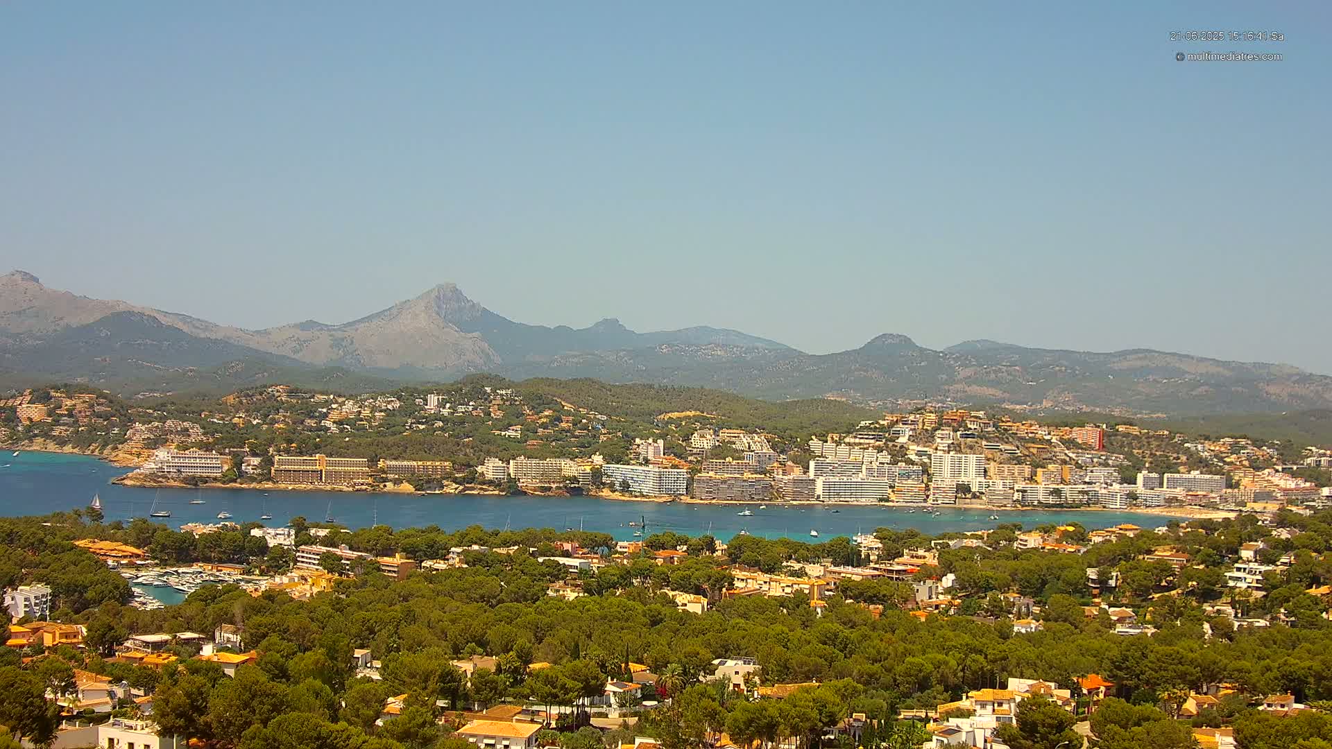 A coastal town nestled among green hills and a calm bay, backed by a mountain range under a clear blue sky.