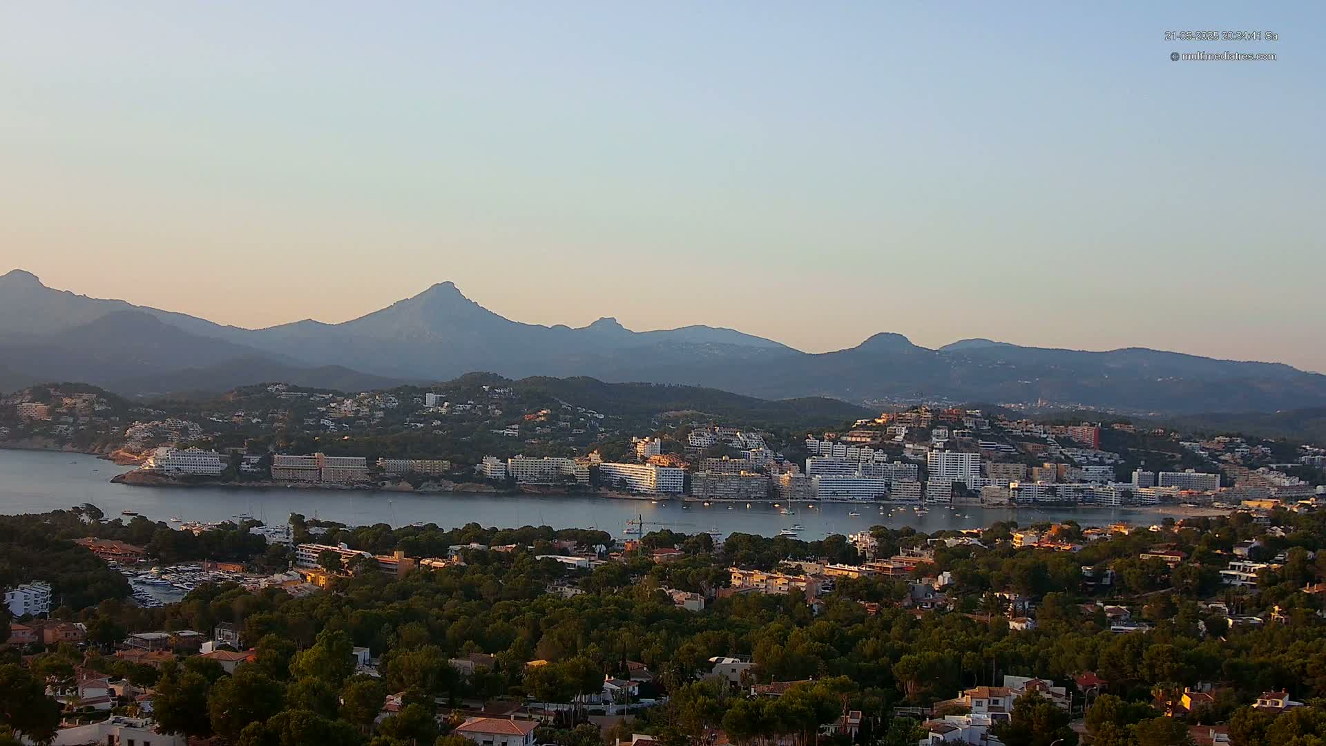 A coastal town nestled amongst green hills and mountains, with calm waters in the foreground under a clear, light sky.