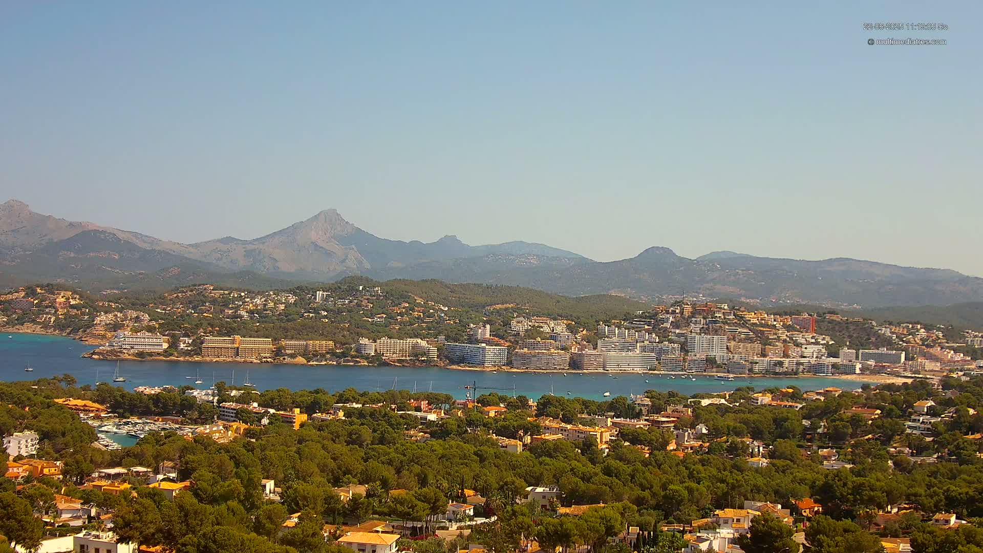 A coastal town nestled amongst green hills and mountains under a clear blue sky, with a calm bay separating the town from the mountains.