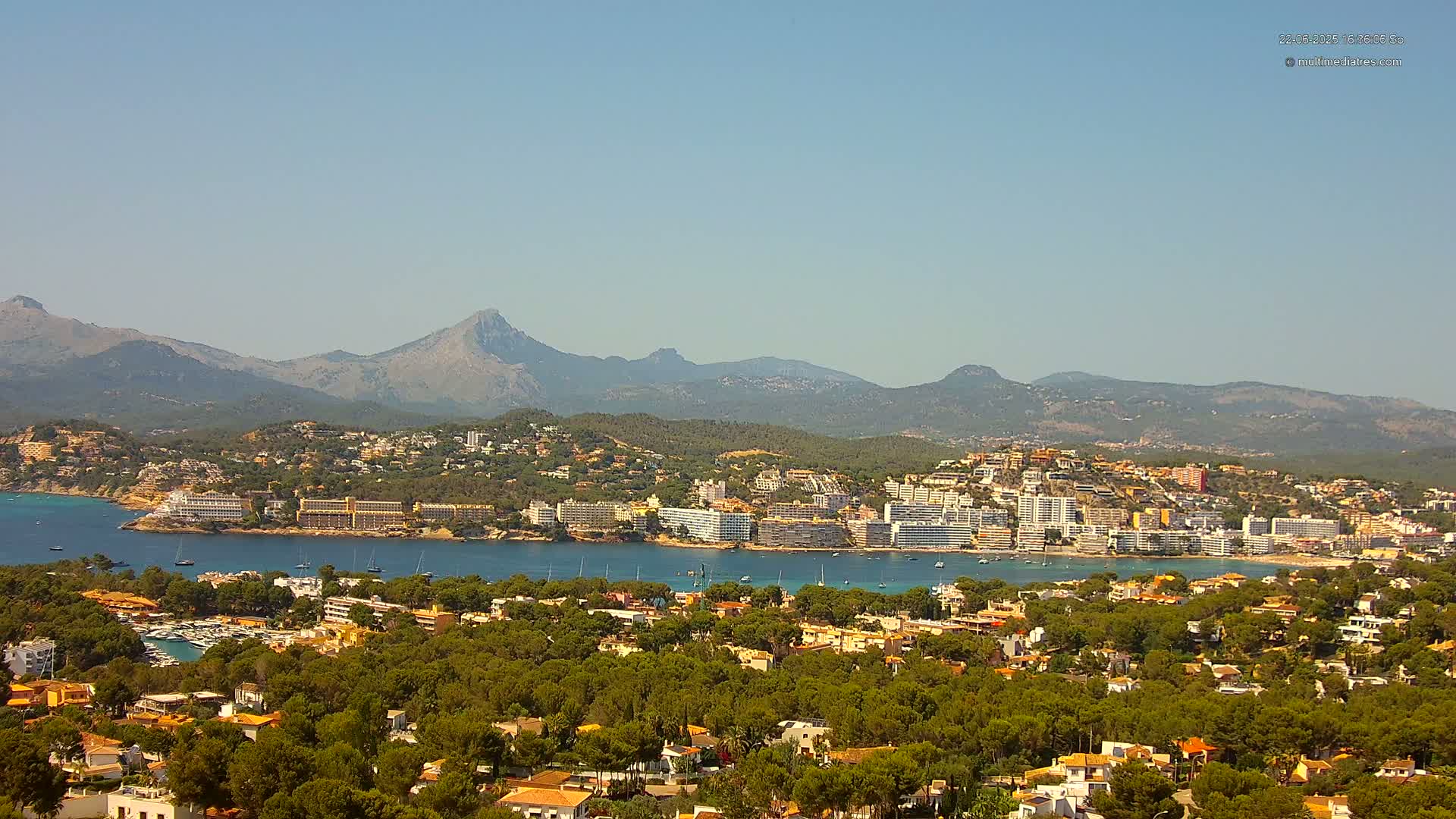 A coastal town nestled amongst green hills and mountains under a clear blue sky, with a calm body of water separating the town from a mountainous backdrop.