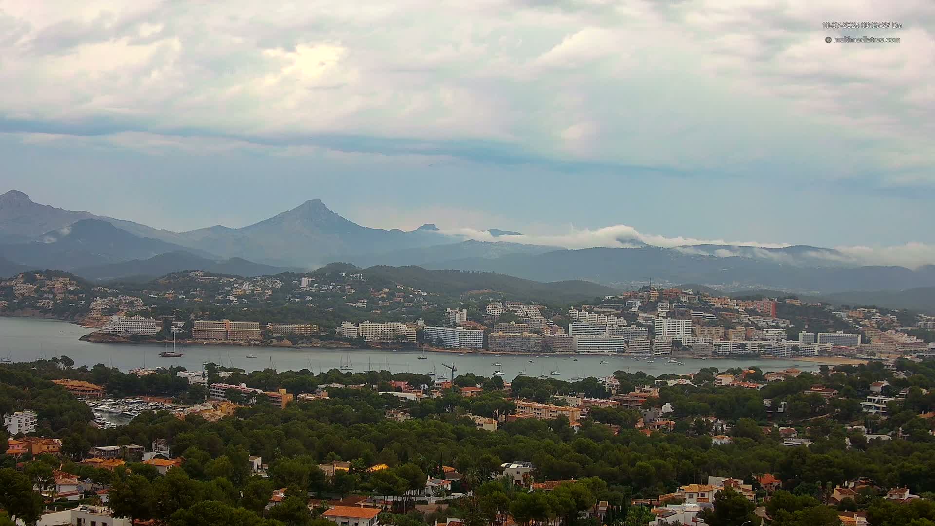 A coastal town nestled amongst green hills and mountains, under a mostly cloudy sky, with a calm bay filled with boats.