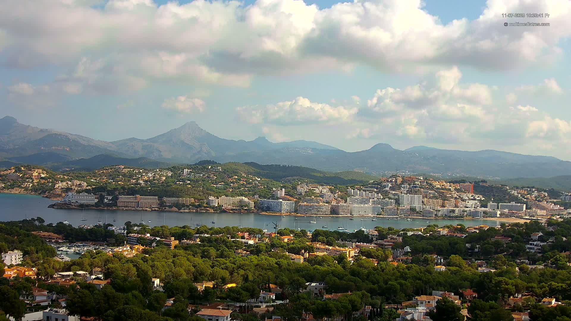 A coastal town nestled between a calm bay and a mountain range under a partly cloudy sky.