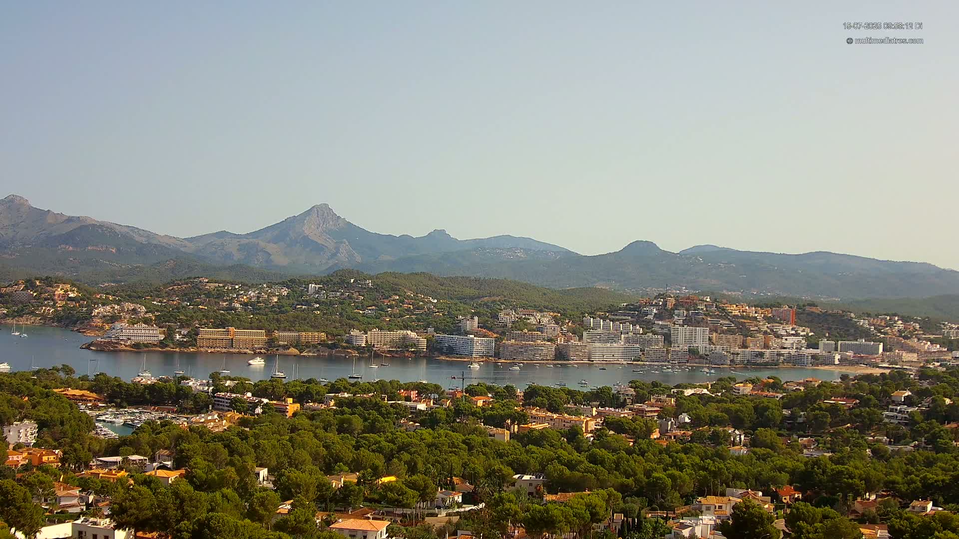 A coastal town nestled amongst green hills and mountains under a clear, sunny sky, with a calm bay filled with boats.