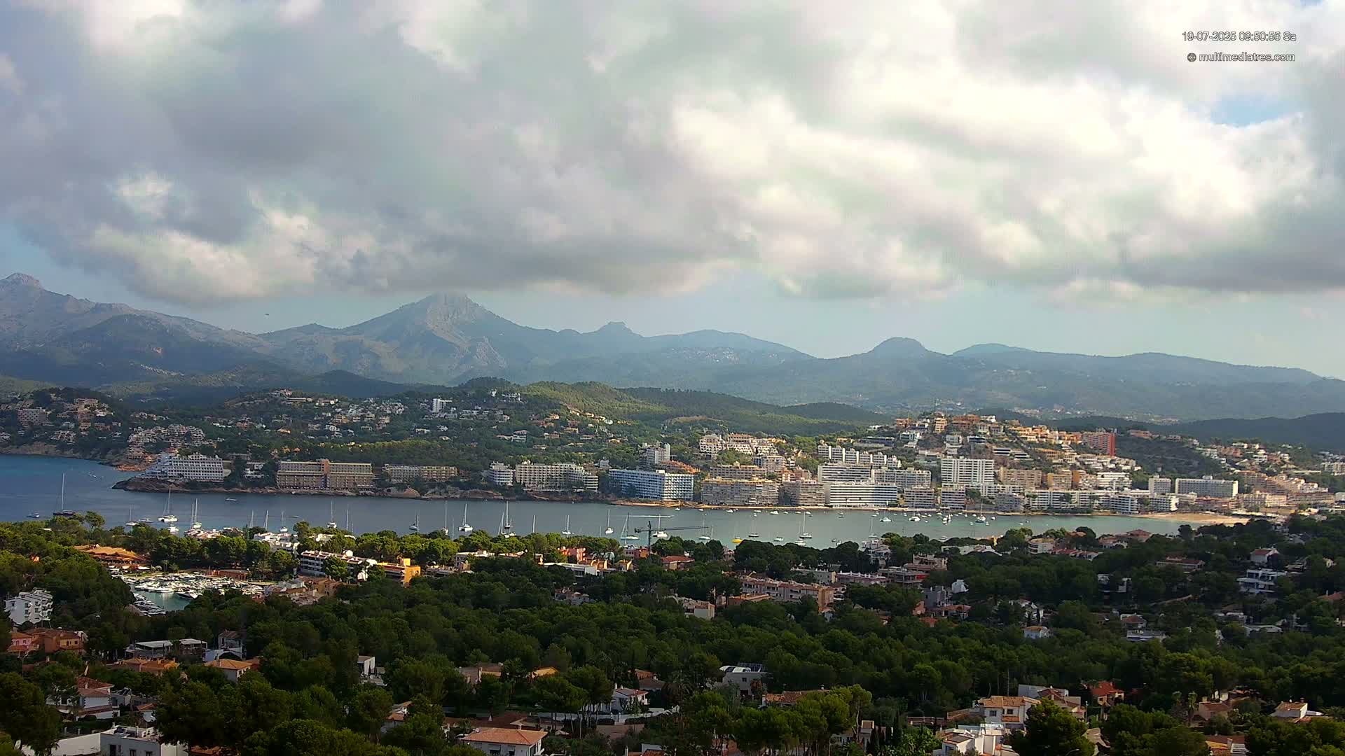 A coastal town nestled between a calm bay and a mountain range under a partly cloudy sky.