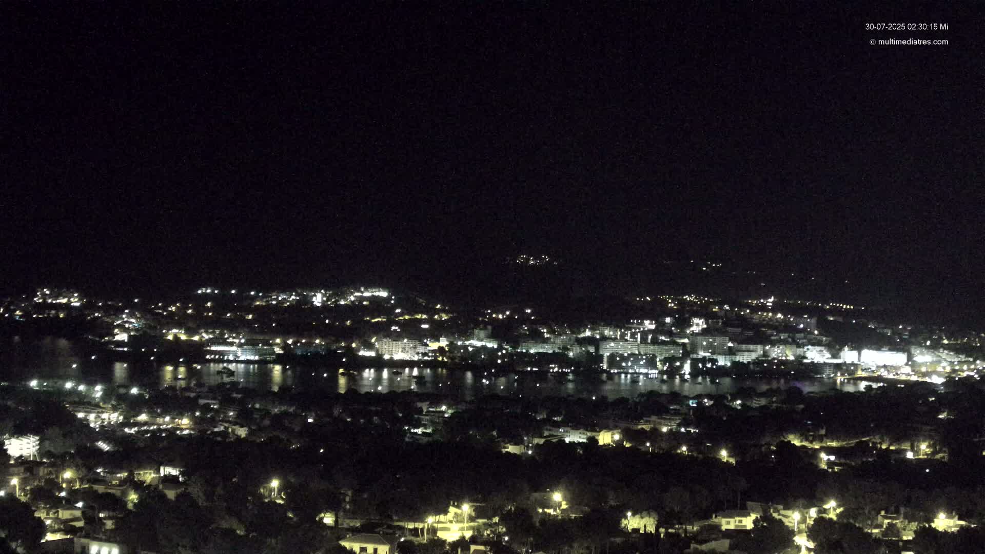A coastal town nestled between a calm bay and a mountain range under a partly cloudy sky.