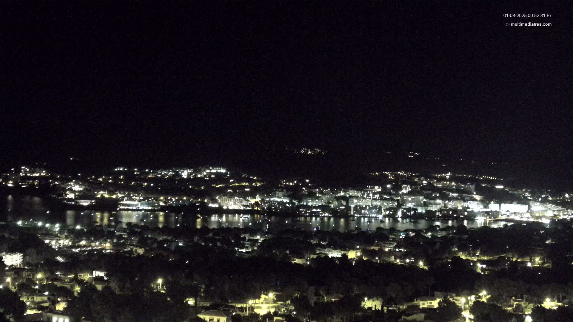 A nighttime, aerial view shows a coastal town illuminated with lights, reflecting on a calm body of water, under a clear, dark sky.