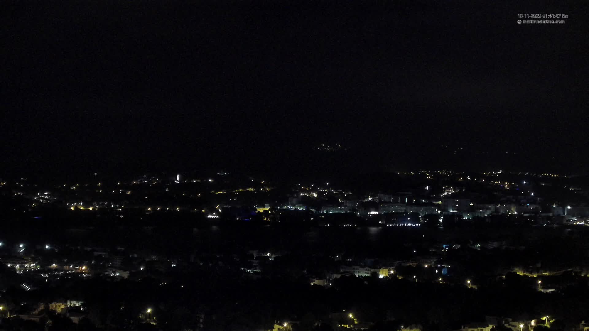 The image captures a panoramic nighttime view of a town or city, illuminated by numerous street and building lights, with a visible body of water reflecting some of the light under a clear, dark sky.