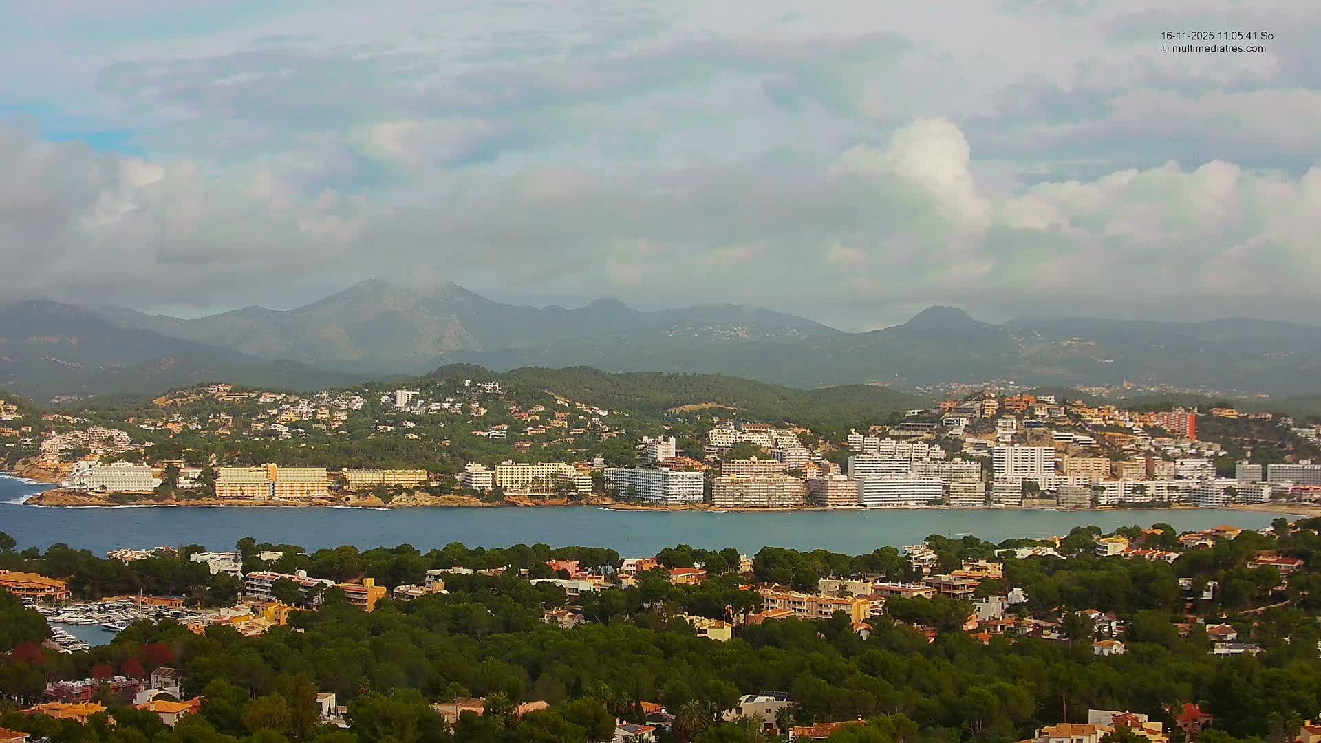 A scenic coastal town featuring densely packed buildings and a marina along a blue bay is framed by tree-covered hills and distant mountains under a partially cloudy sky.