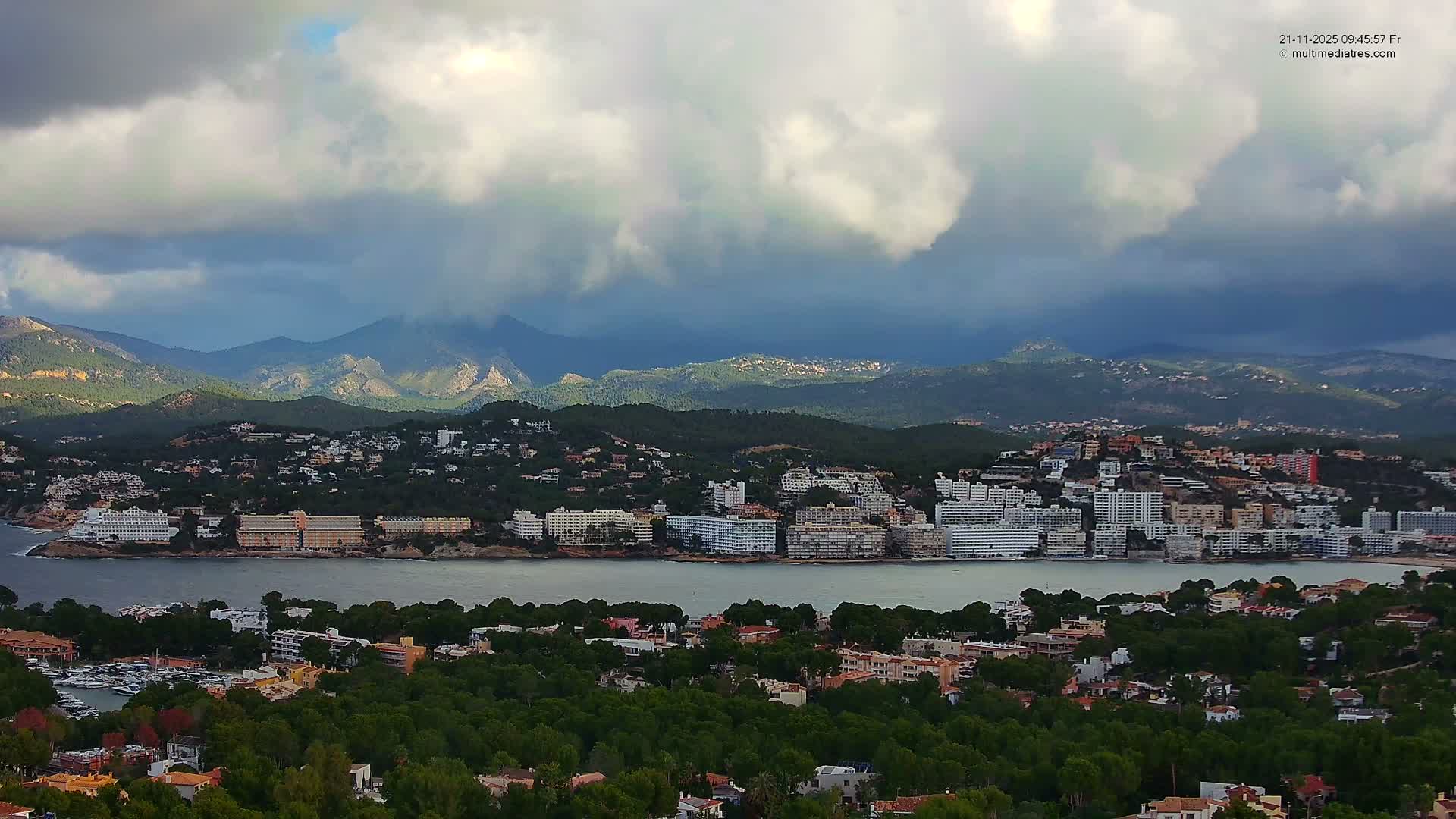 A sprawling coastal town featuring numerous waterfront buildings and a marina is nestled along a bay, flanked by green hills and distant mountains under a dramatic, heavily clouded sky with sporadic patches of brightness.