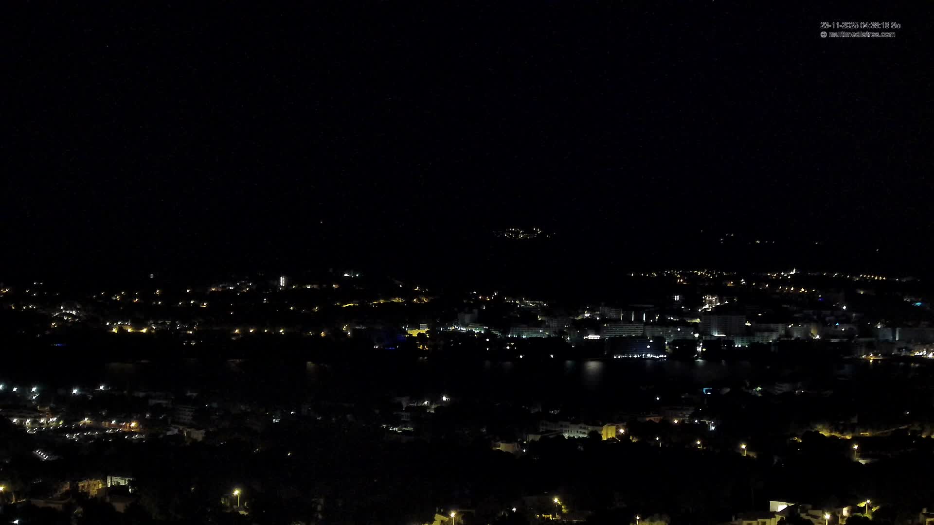 A panoramic nighttime view reveals a sprawling coastal town illuminated by countless lights, bordering a dark body of water under a clear night sky.