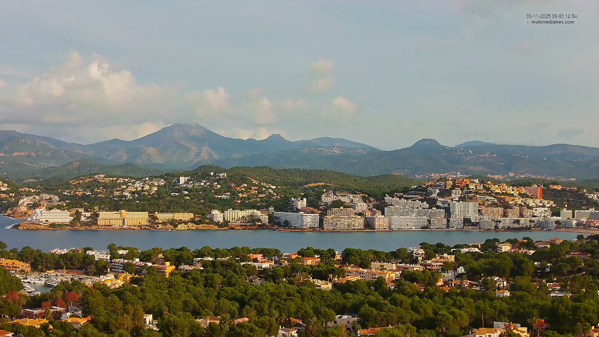 A scenic coastal town sprawls around a calm bay, with buildings climbing up tree-covered hills and mountains rising in the distance under a bright sky with scattered clouds.