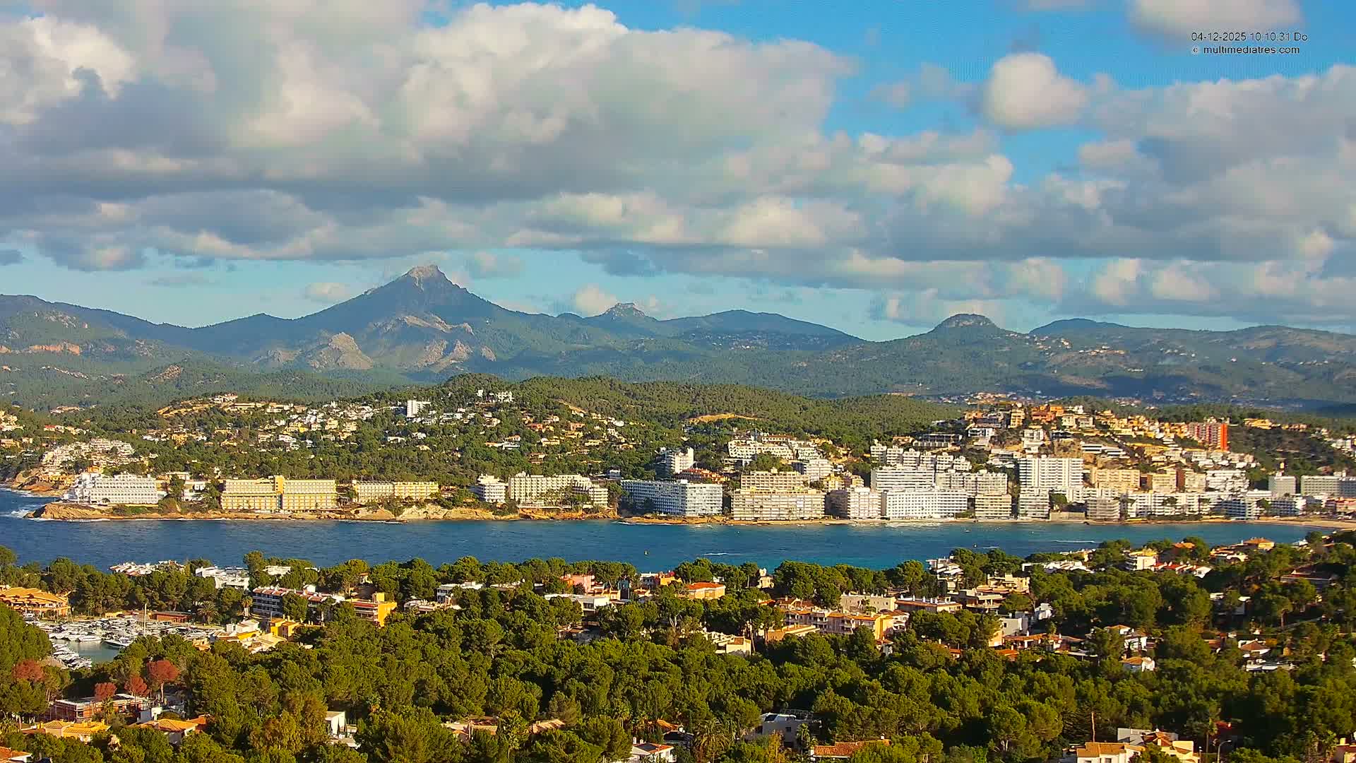 A sunny coastal landscape features a town with many buildings along a vivid blue bay, flanked by tree-covered hills in the foreground and majestic mountains in the distance, all beneath a bright blue sky with scattered white clouds.