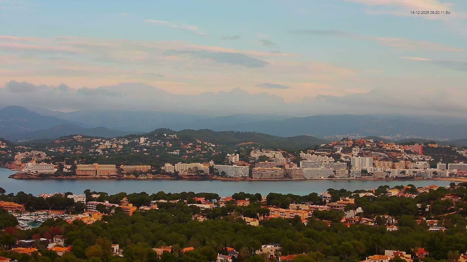 A sunny coastal landscape features a town with many buildings along a vivid blue bay, flanked by tree-covered hills in the foreground and majestic mountains in the distance, all beneath a bright blue sky with scattered white clouds.