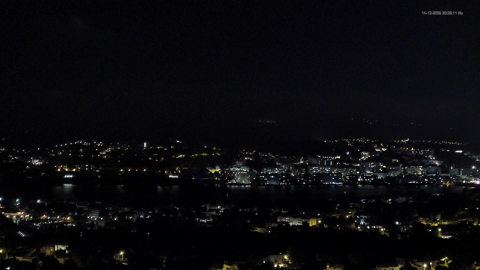 A sunny coastal landscape features a town with many buildings along a vivid blue bay, flanked by tree-covered hills in the foreground and majestic mountains in the distance, all beneath a bright blue sky with scattered white clouds.