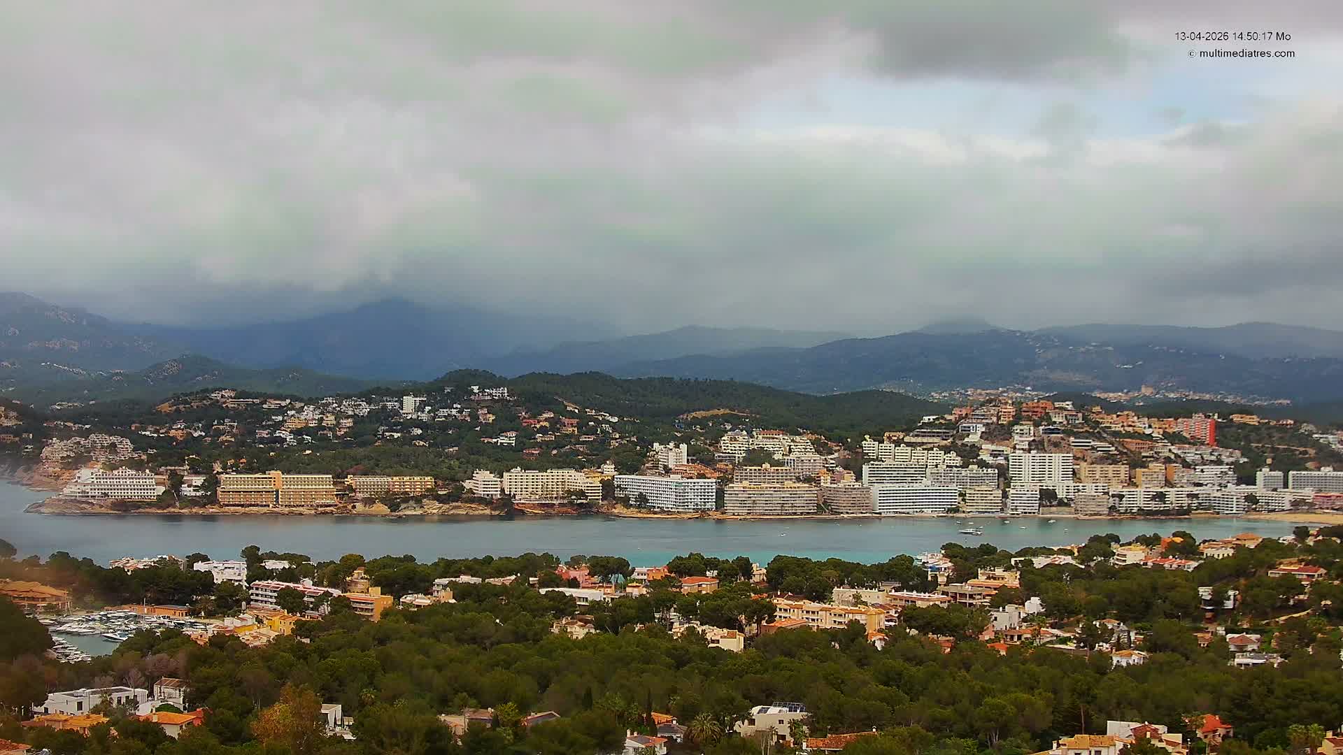 A sunny coastal landscape features a town with many buildings along a vivid blue bay, flanked by tree-covered hills in the foreground and majestic mountains in the distance, all beneath a bright blue sky with scattered white clouds.