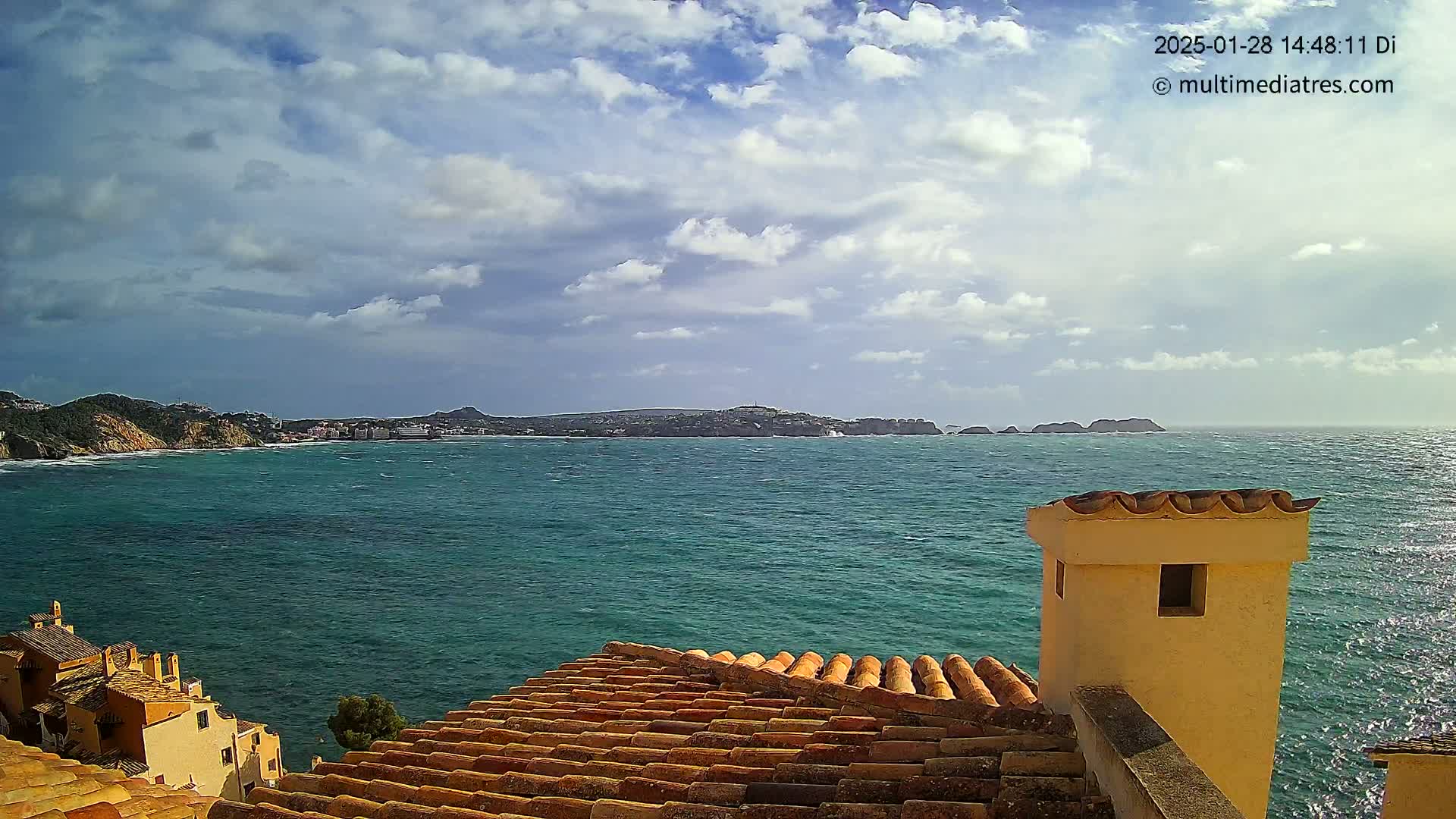 A partly cloudy sky overlooks a teal ocean with distant landmasses, viewed from a terracotta-tiled roof with a small, light-yellow chimney.