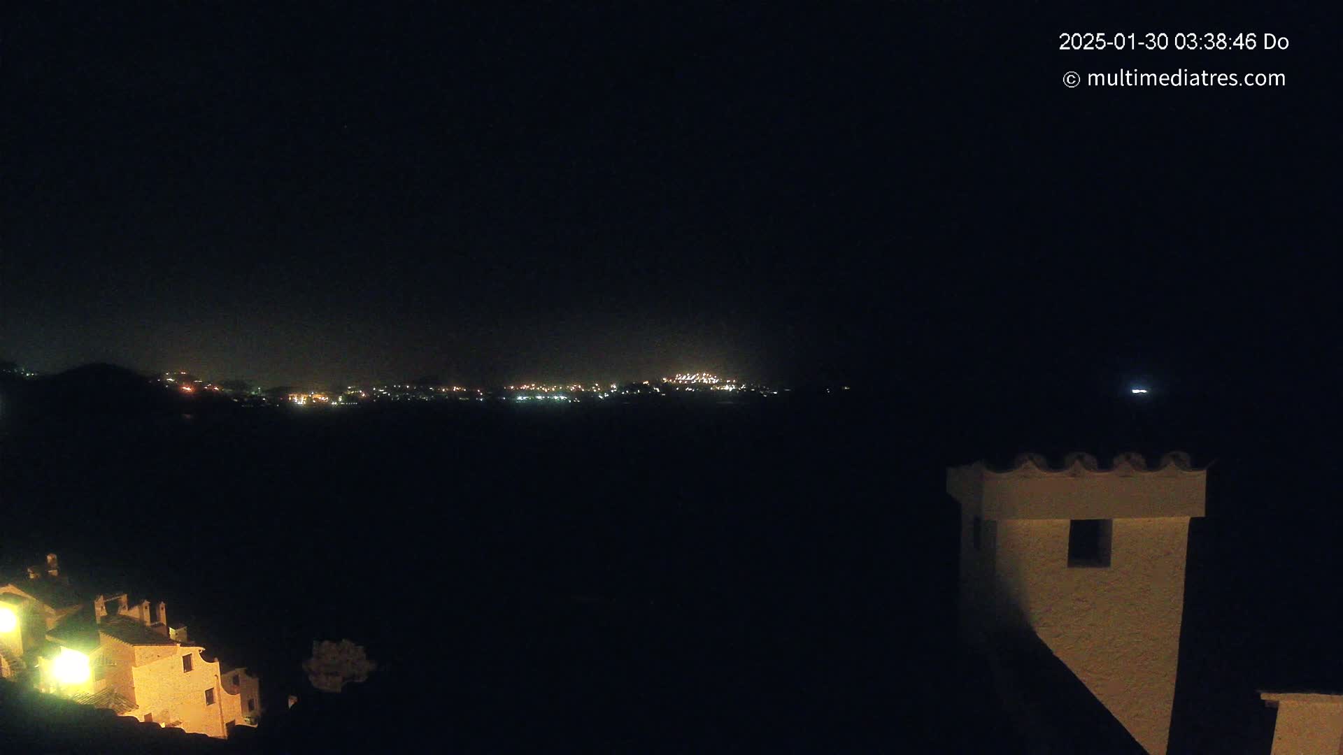 A nighttime view shows a distant town illuminated with lights across a dark valley, observed from a vantage point near a building with a distinctive chimney.