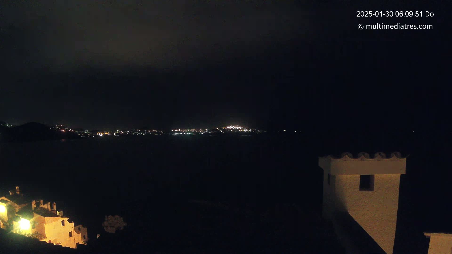 A nighttime view shows a dark body of water separating a dimly lit coastal town from a building with a crenellated top, under a cloudy, dark sky.
