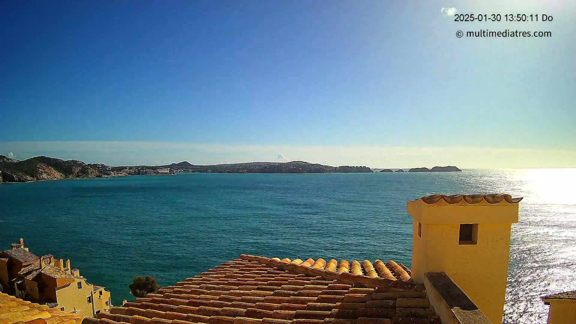 A terracotta roof overlooks a calm, teal ocean with distant hills under a clear blue sky.