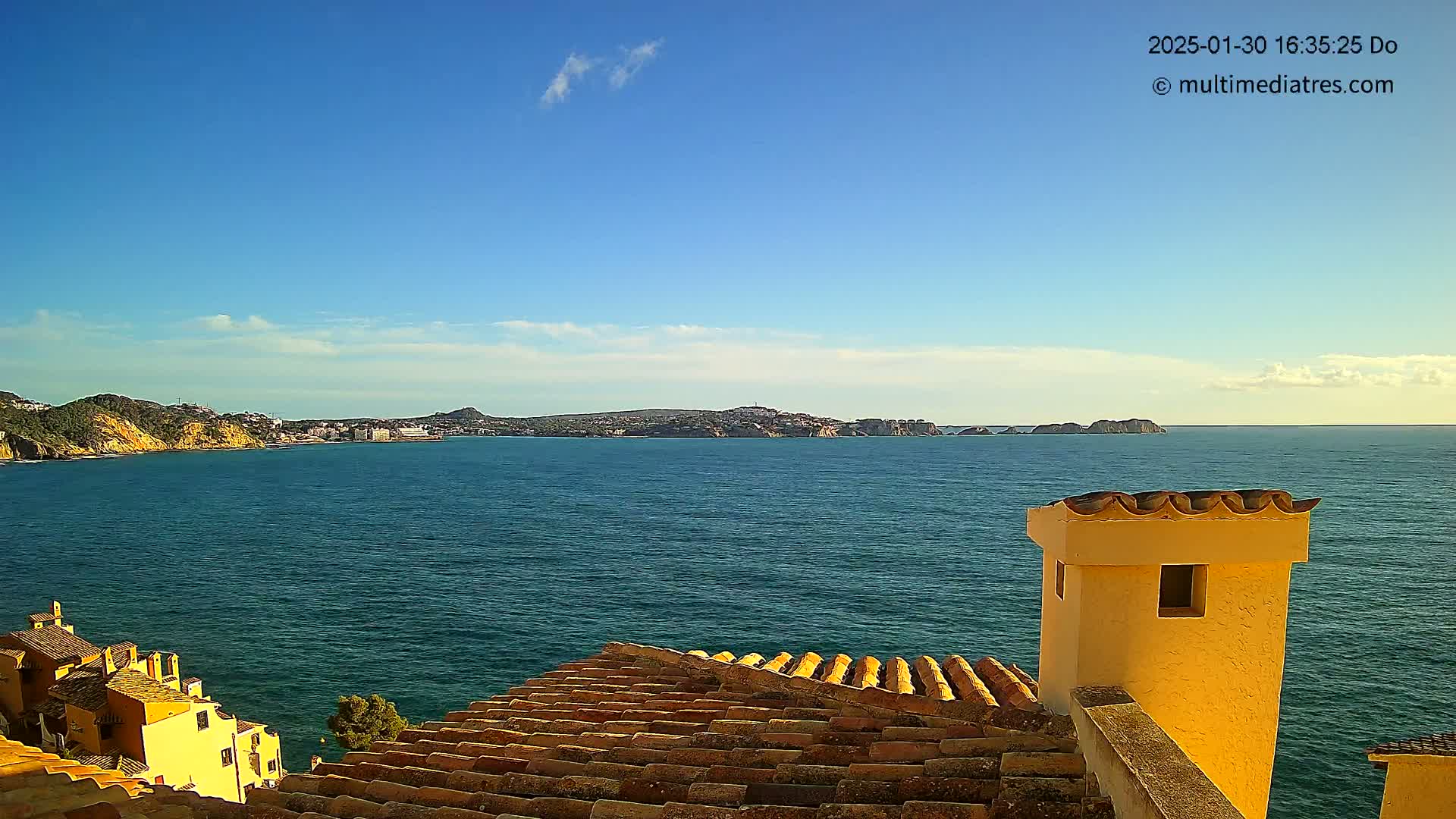 A coastal view on a sunny day shows a calm blue sea stretching to a distant coastline, partially obscured by a tiled roof and a yellow chimney in the foreground.