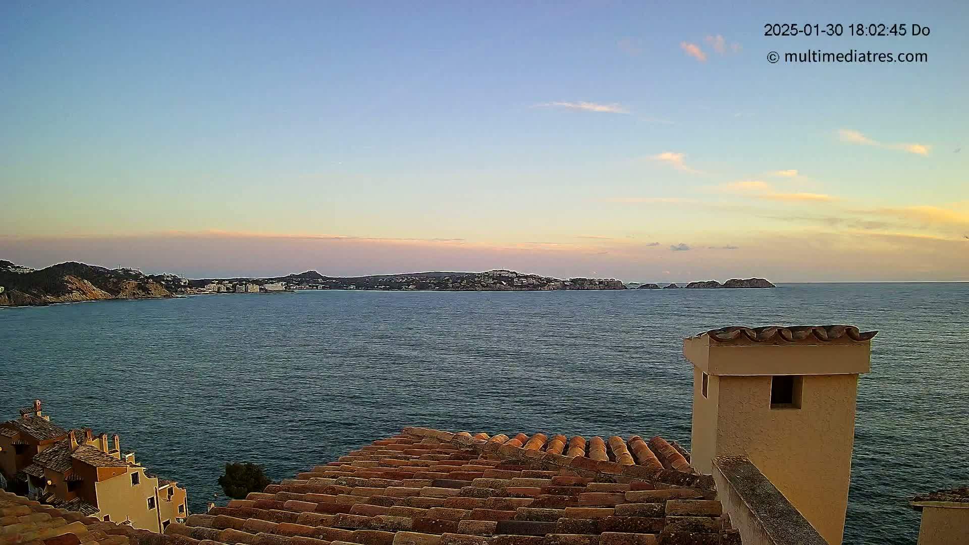 A coastal scene at sunset shows a calm sea with a distant island and shoreline, viewed from a rooftop with terracotta tiles and a light-beige chimney.