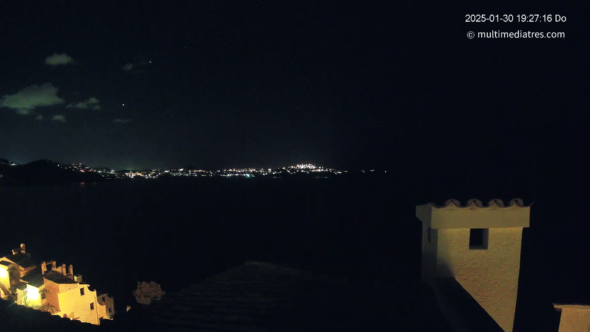 A nighttime view of a city skyline across a dark body of water, partially obscured by clouds, with a portion of a building and chimney in the foreground.