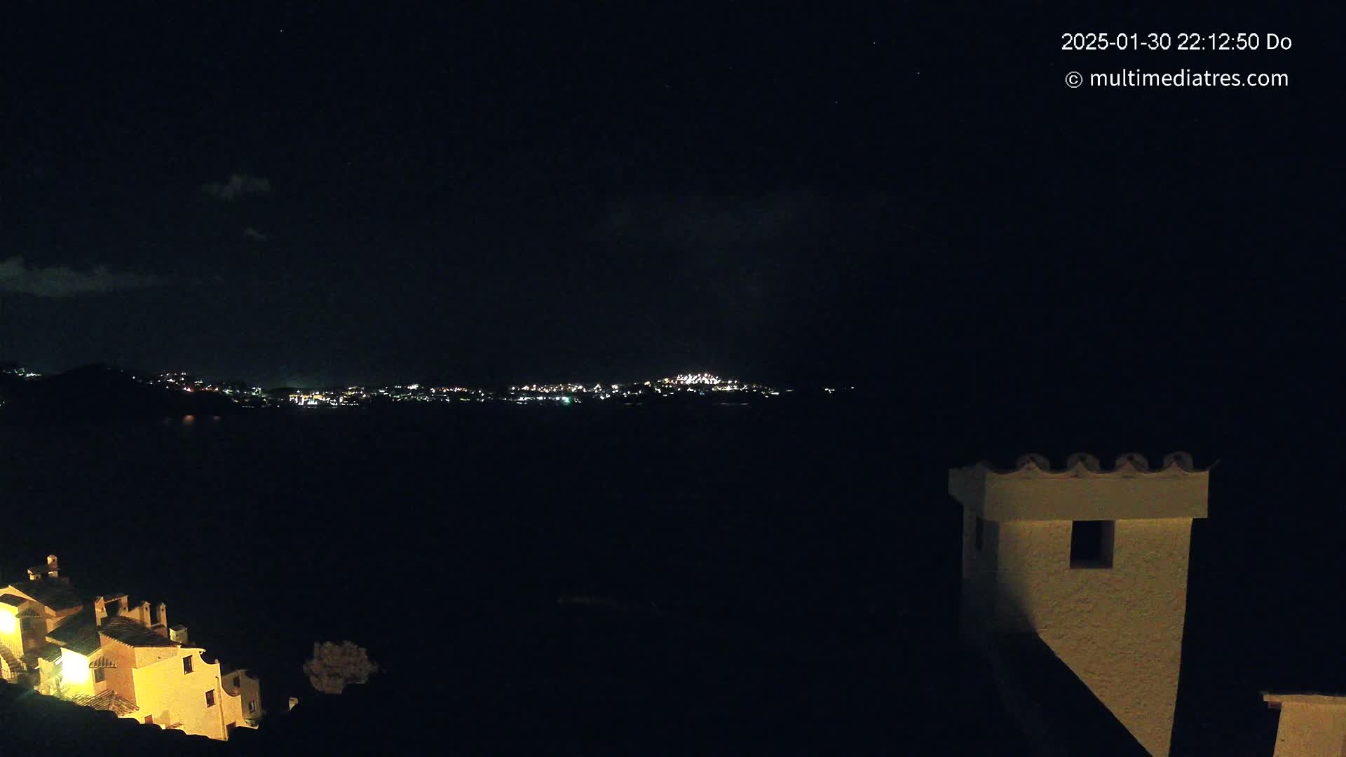 A nighttime view of a city's lights across a dark body of water, partially obscured by hills, with a tan building and its chimney in the foreground under a clear, starry sky.
