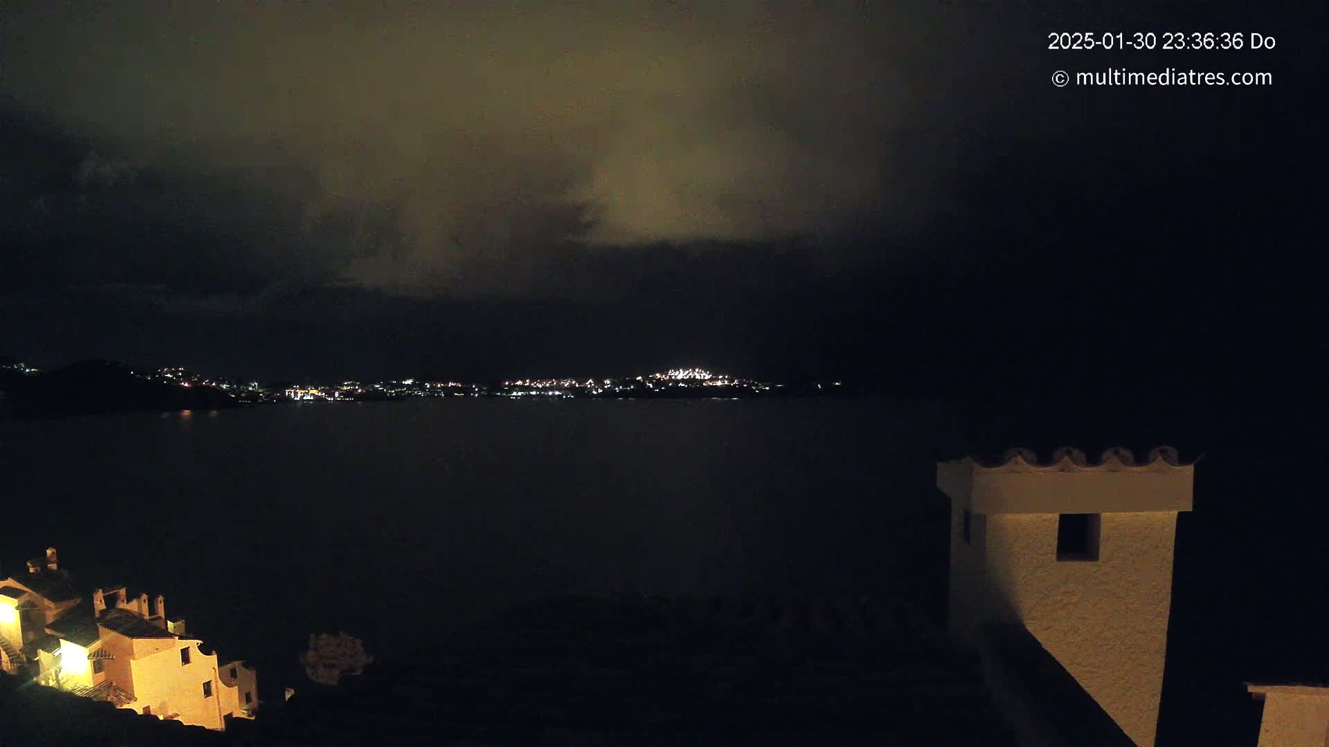 A nighttime view of a city across a dark body of water, under a cloudy sky, with illuminated buildings in the foreground.