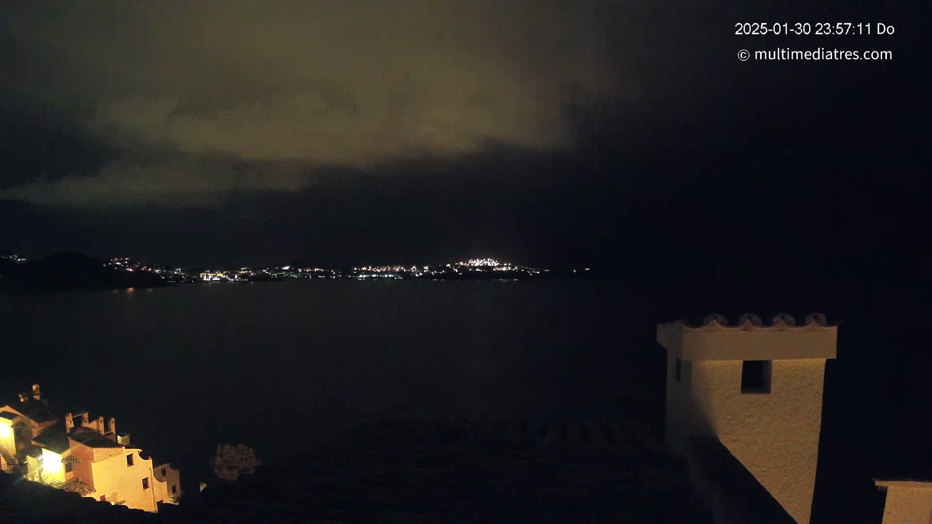 A nighttime view of a dark body of water with a city's lights across it, under a cloudy sky, with buildings and a chimney visible in the foreground.