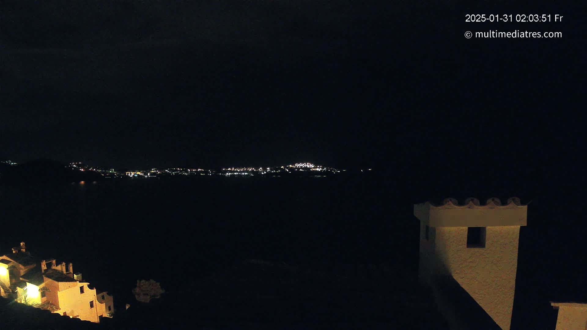 A nighttime view of a city's lights across a dark body of water, with a portion of a building in the foreground.