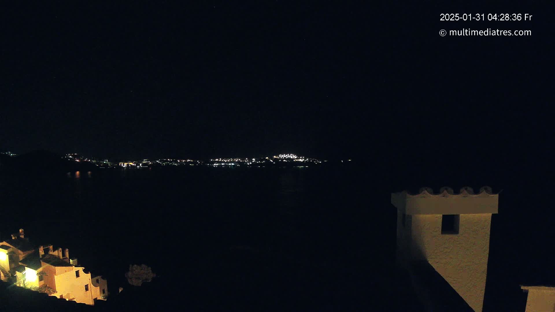 A nighttime view of a city's lights across a dark body of water, with a building partially visible in the foreground.
