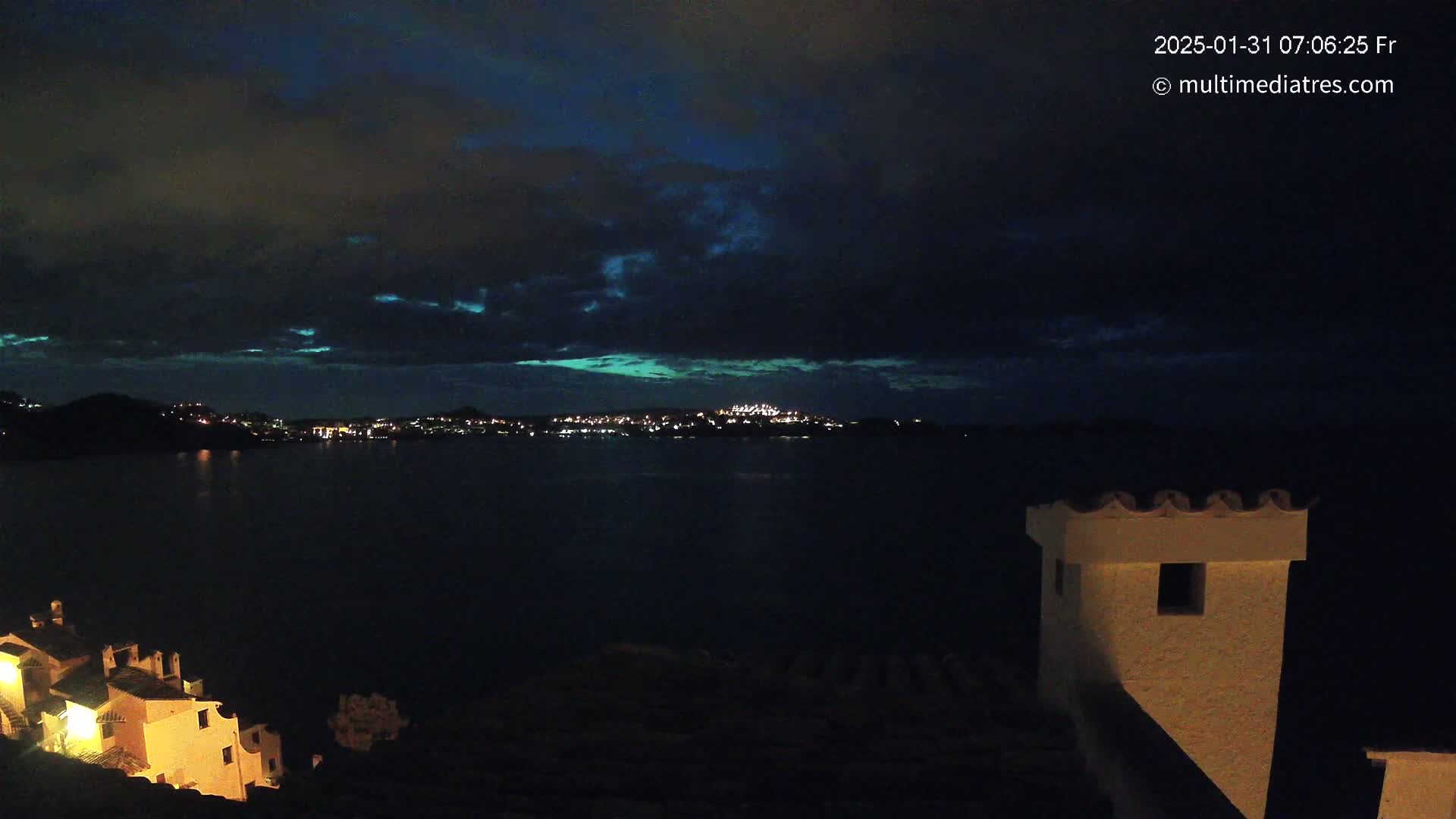 A nighttime view of a town's lights across a dark body of water, under a partly cloudy sky.